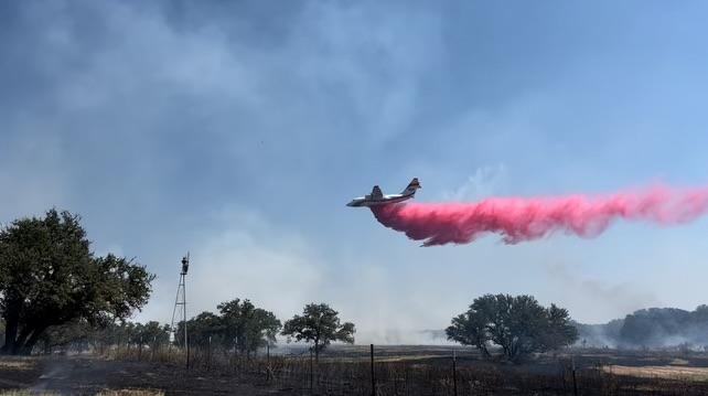 Large airtanker dropping red fire retardant on the North Art Complex fire. Small fires and a windmill can be seen on the left-hand side of the photo. Smoke can be seen in the sky and closer towards to ground at the bottom of the photo.