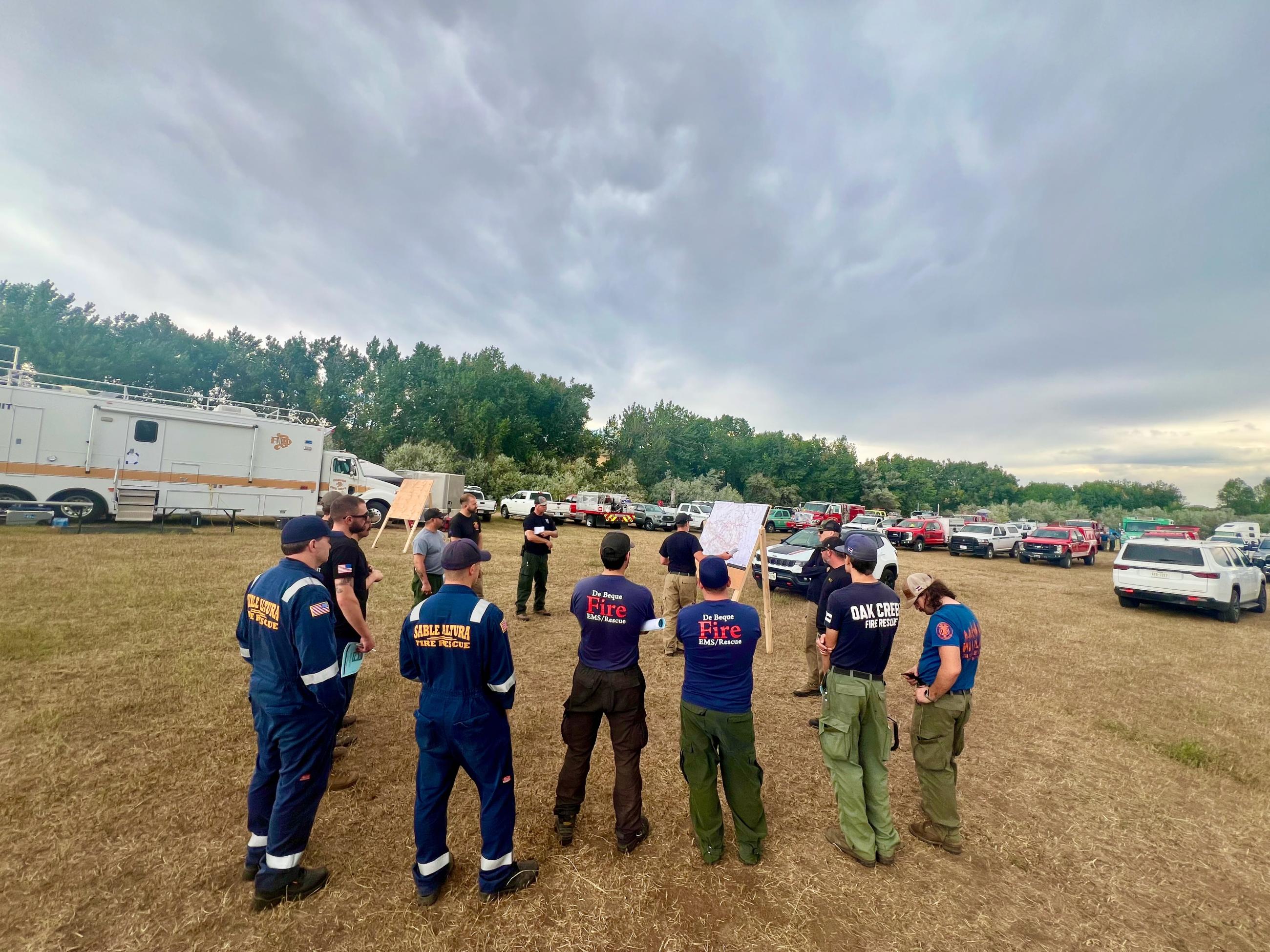 A group of fire personnel stand in a semi-circle around their supervisor standing next to a map on a sandwich board. Low grey clouds fill the upper third of the picture.