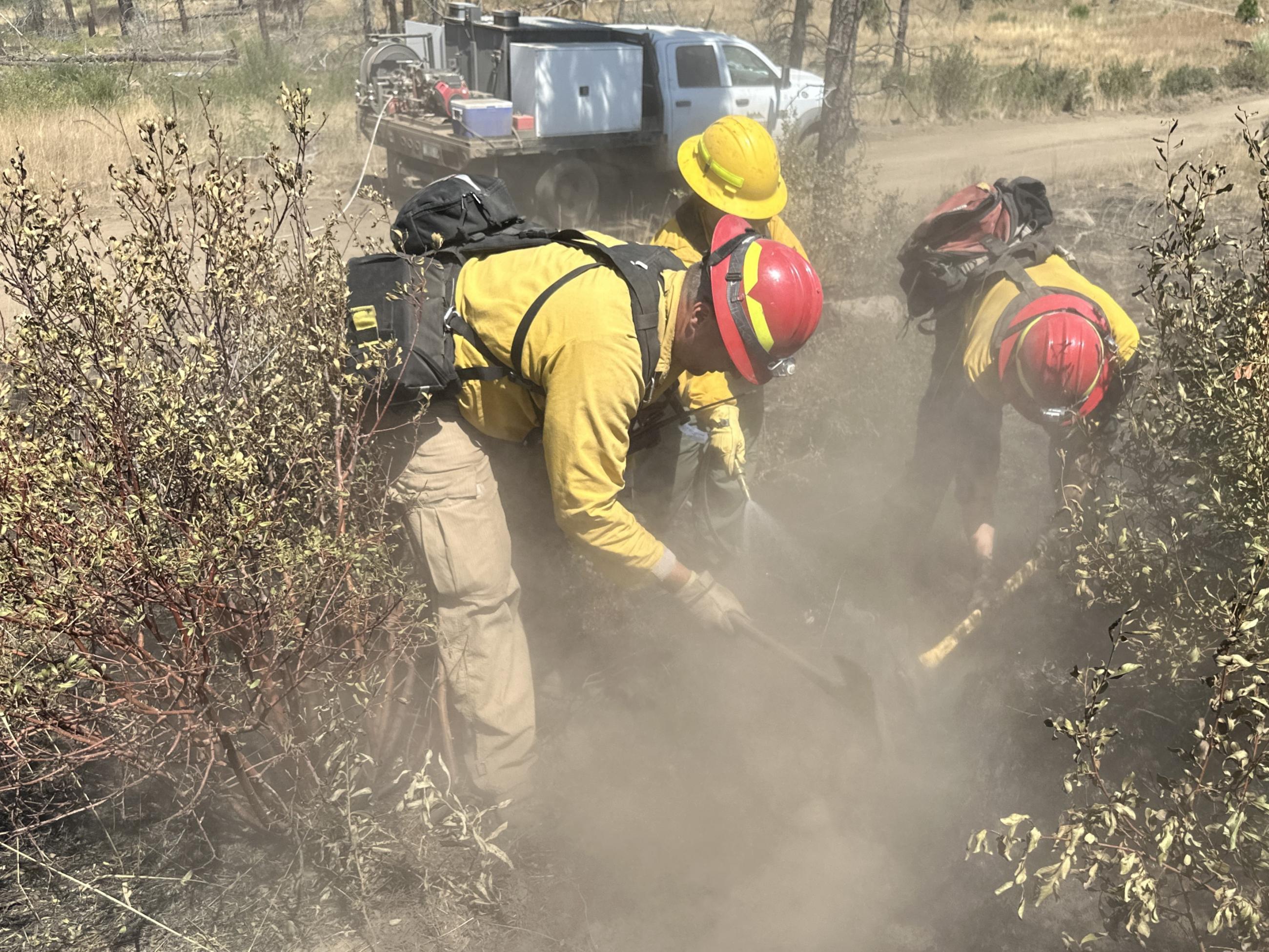 Three firefighters wearing personal protective equipment spray water and use hand tools in a dry brushy area working up a cloud of dust