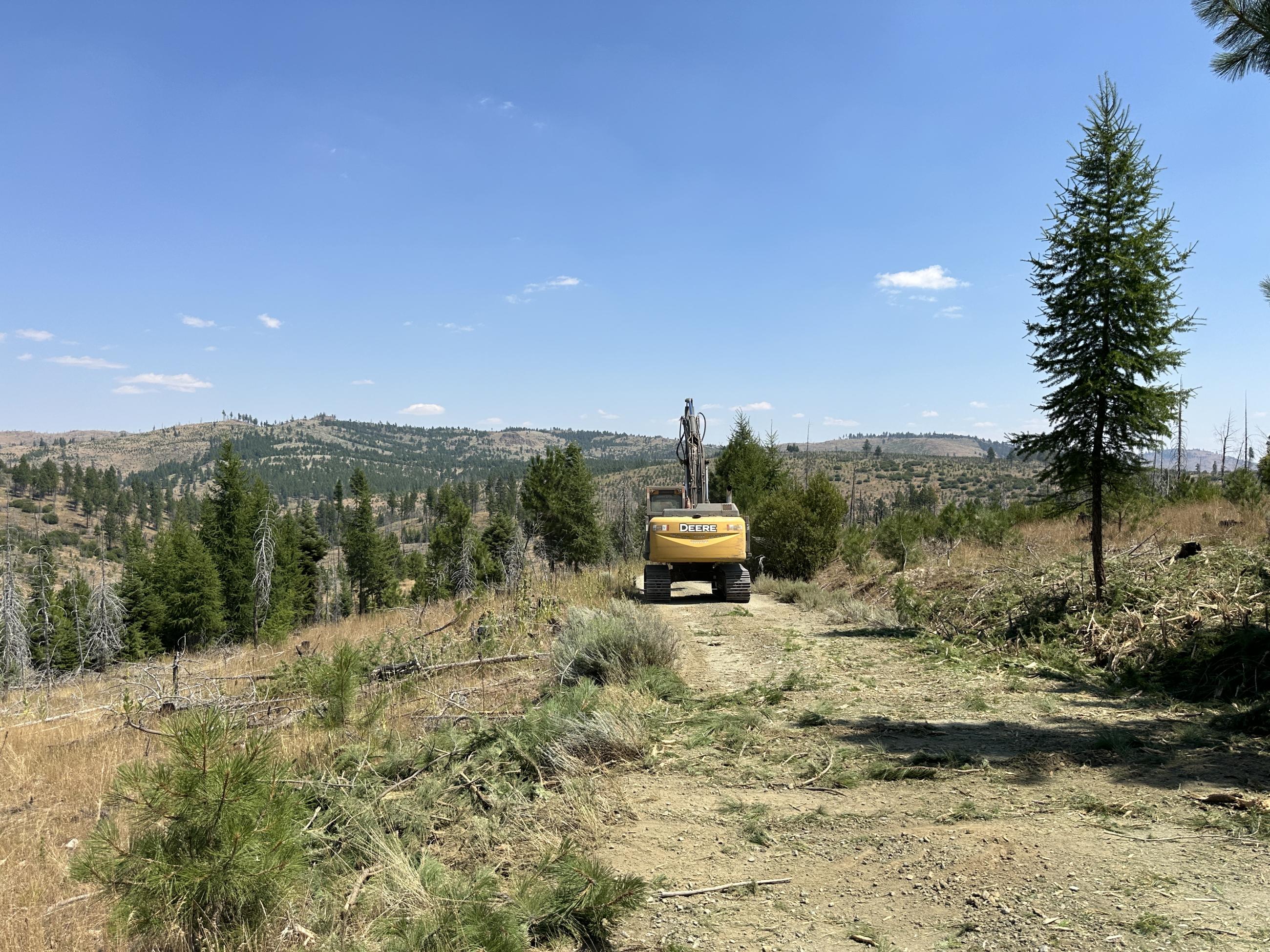 A tractor type machine drives down the middle of a dirt road surrounded by brushy hillsides with scattered trees