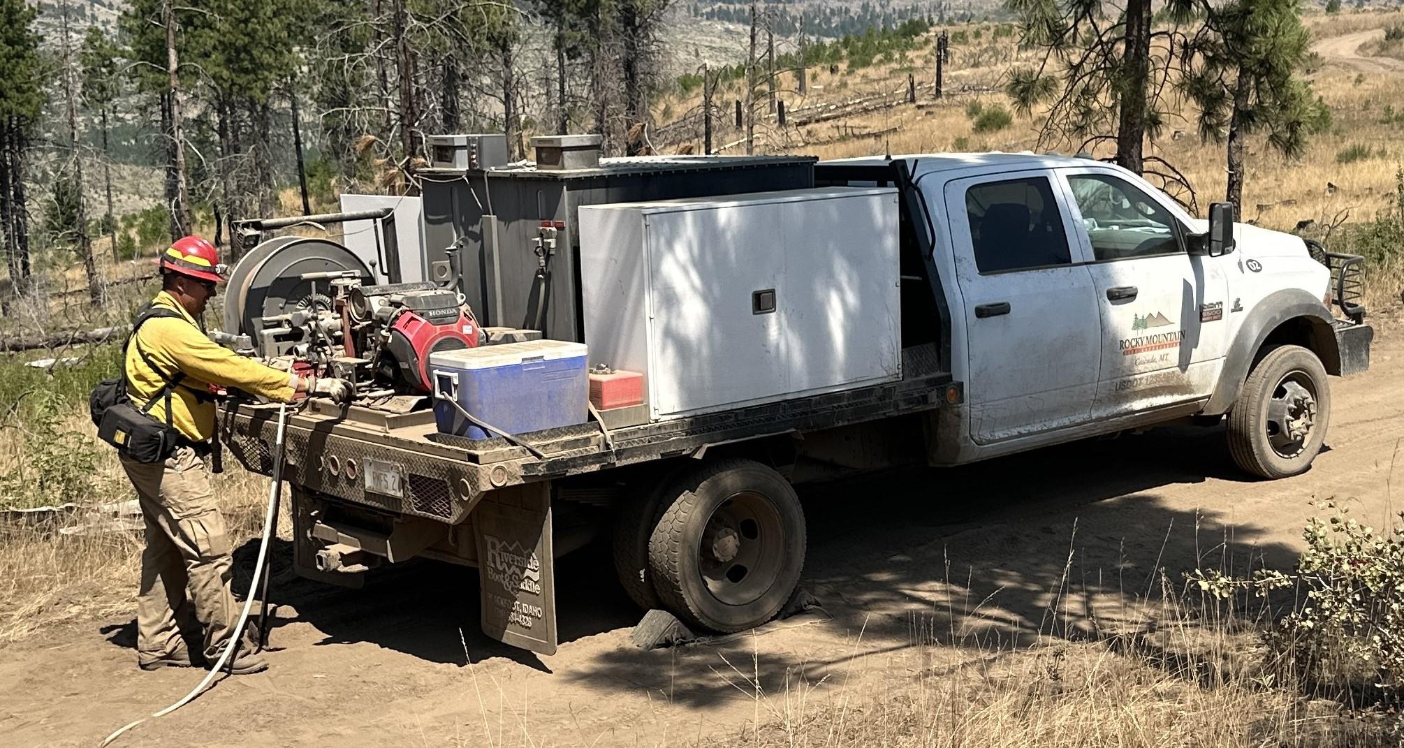 Firefighter wearing personal protective equipment stands at the back of a wildland fire engine with mechanical equipment loaded onto flatbed