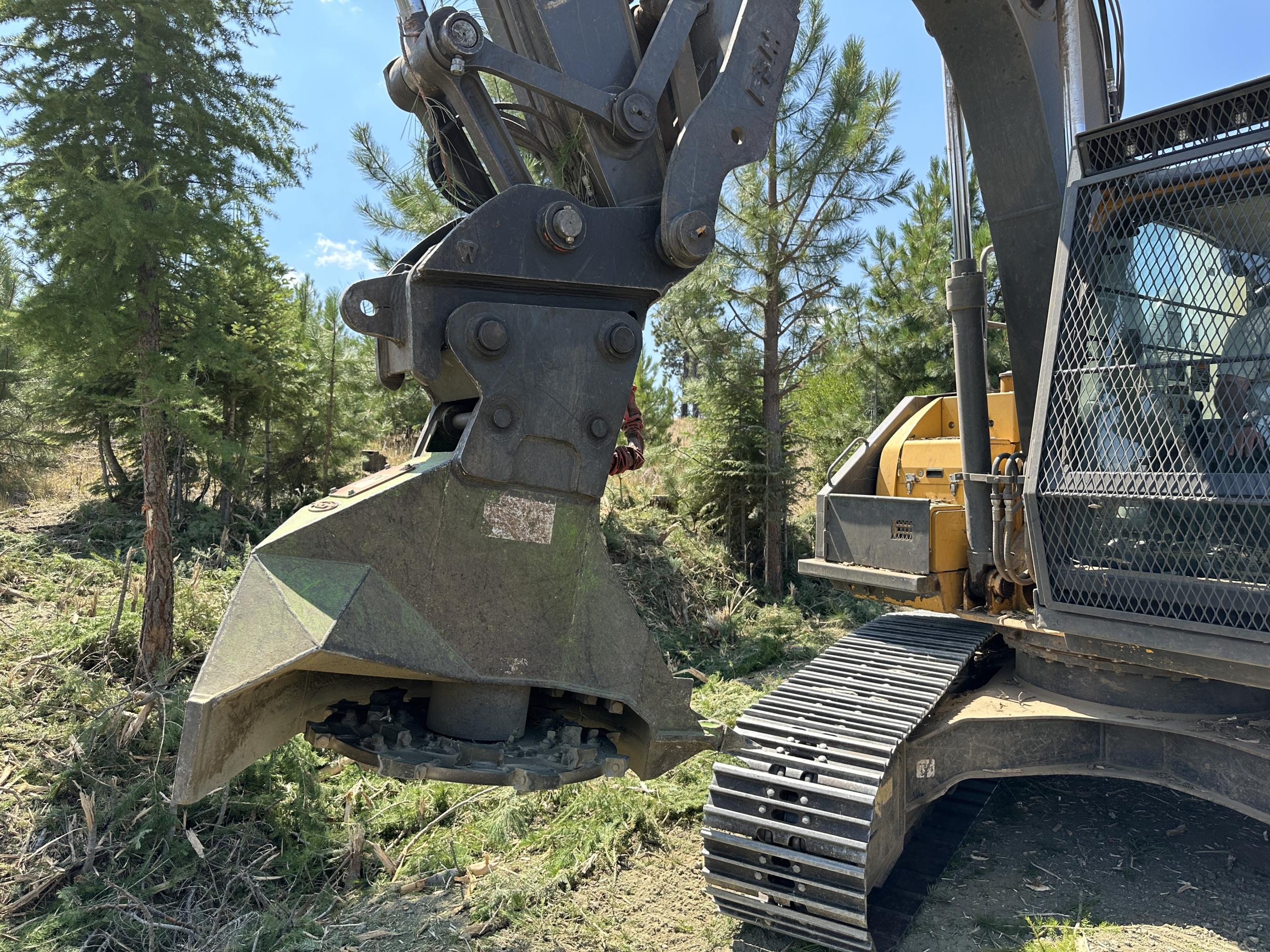 Grinding tool attached to tractor type machinery on a dirt road next to a field of grass and trees