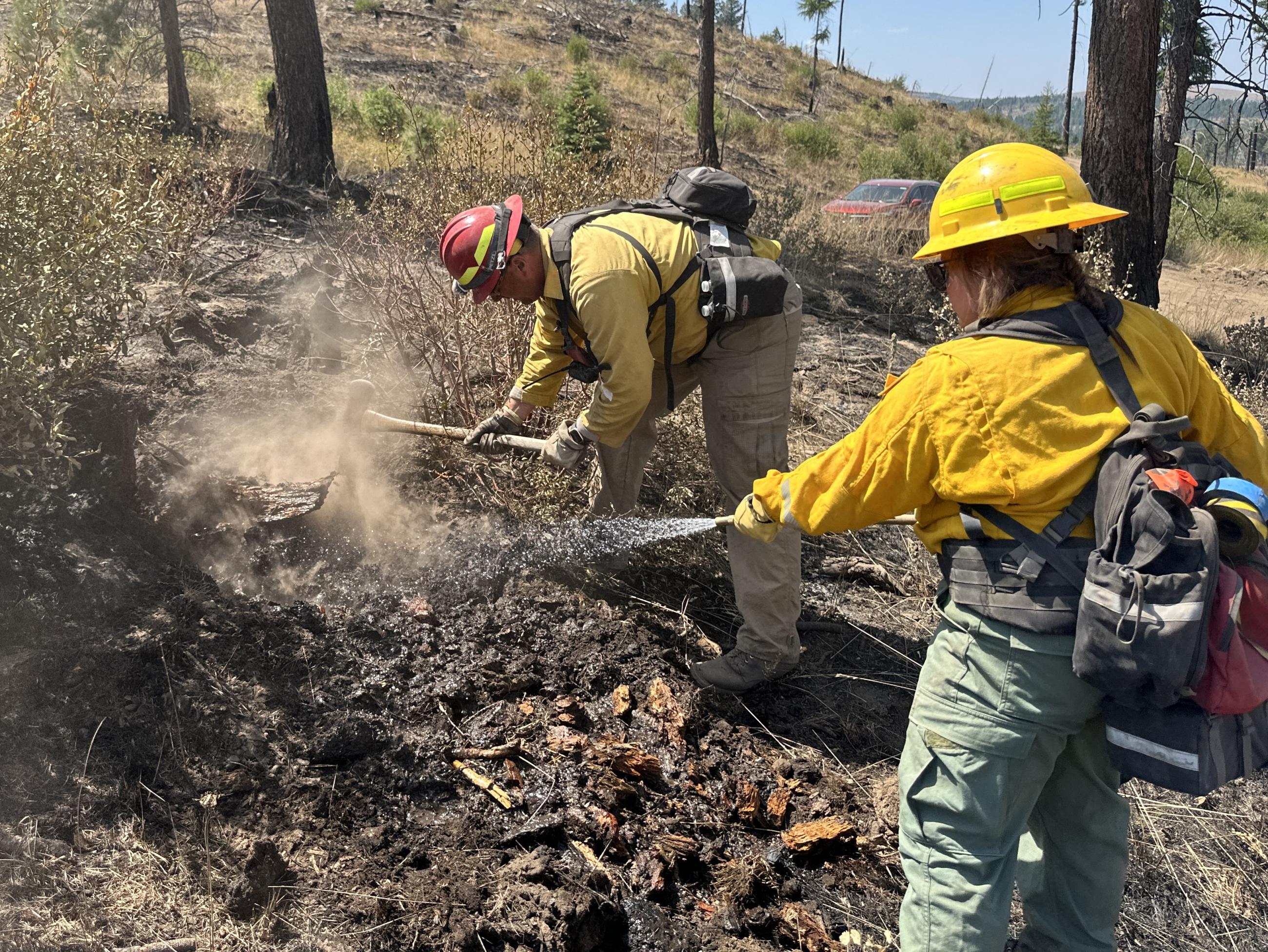 Firefighter in personal protective equipment sprays water from a hose while a second firefighter digs in the dirt raising a cloud of dust on a dry brushy hillside