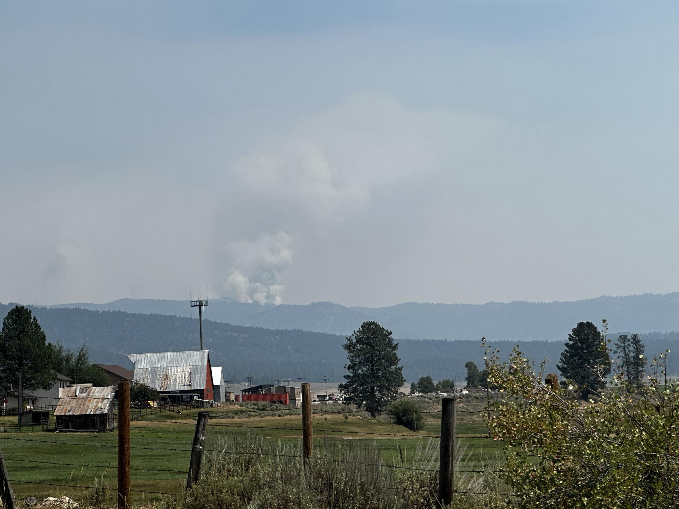 Photo shows a smoke column rising in the up over the mountain top with structures in the foreground