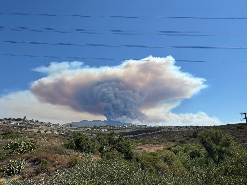 A smoke column rises up and spreads in to a flattened broad head in front of mountains. Clear blue sky behind the column is visible above. Photo taken from a good distance from the fire. 