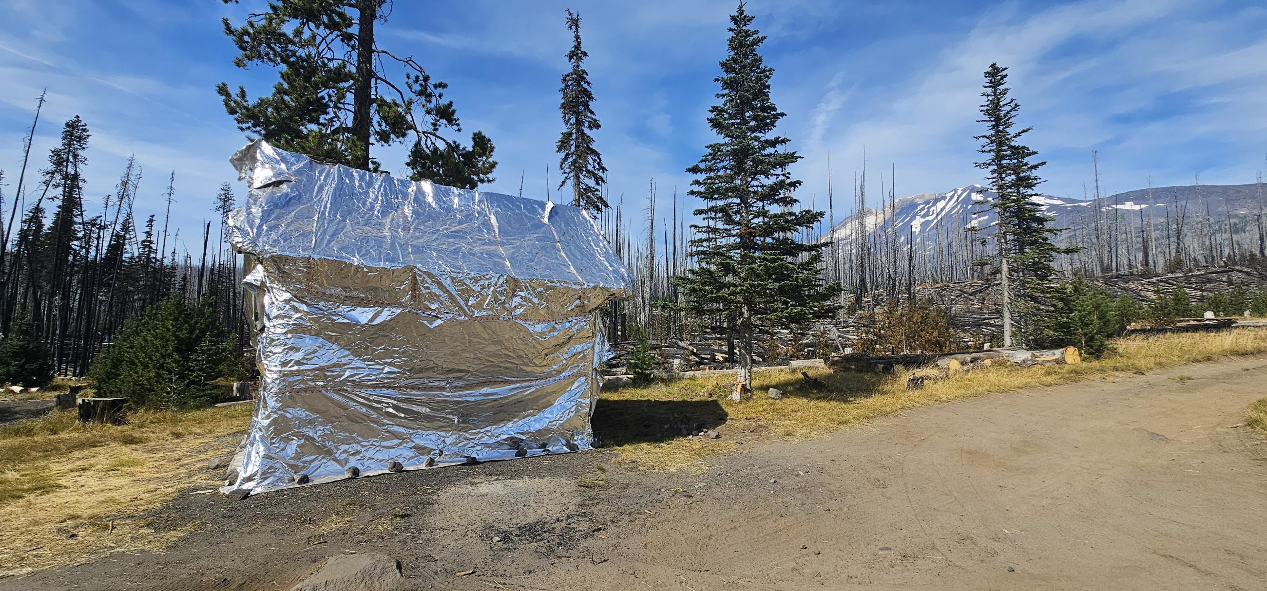 The Morrison Shelter, wrapped in silver-colored protective fabric. The area immediately adjacent is still green. In the background, the ground under the snags is blackened.