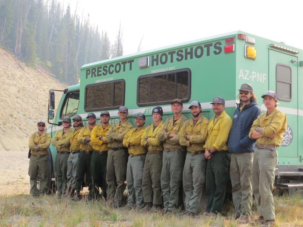 Several firefighters in dirty yellow shirts and dirty green pants pose standing in front of a green wildland fire engine