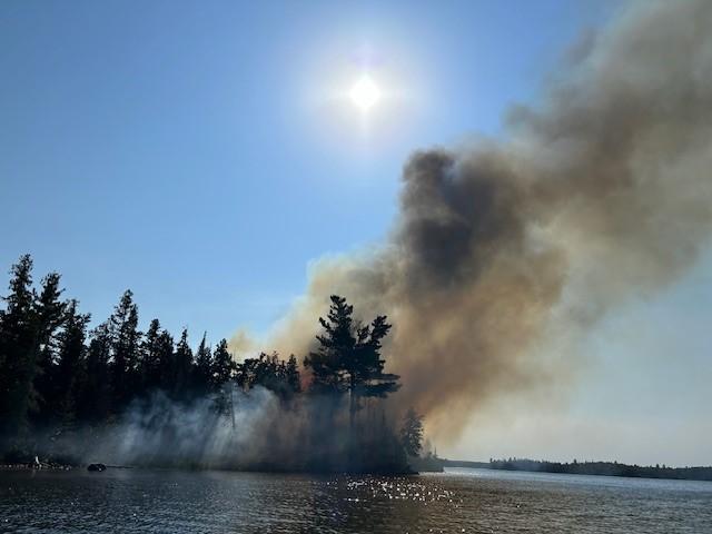 Dark smoke billows into the sky from a wildfire burning on a land point into a lake. 