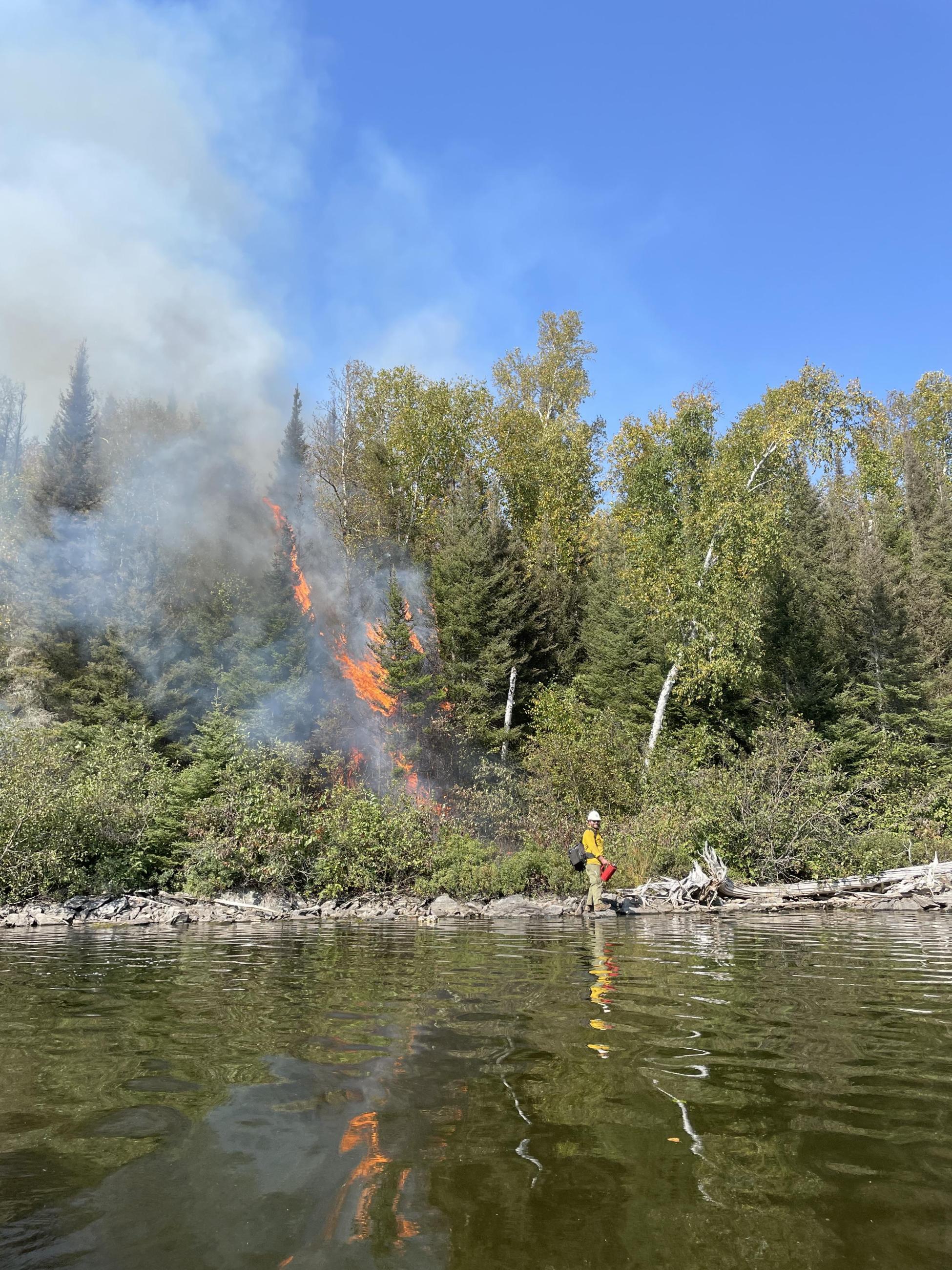 Image of a firefighter on the shoreline of a wildfire on land by a lake. 