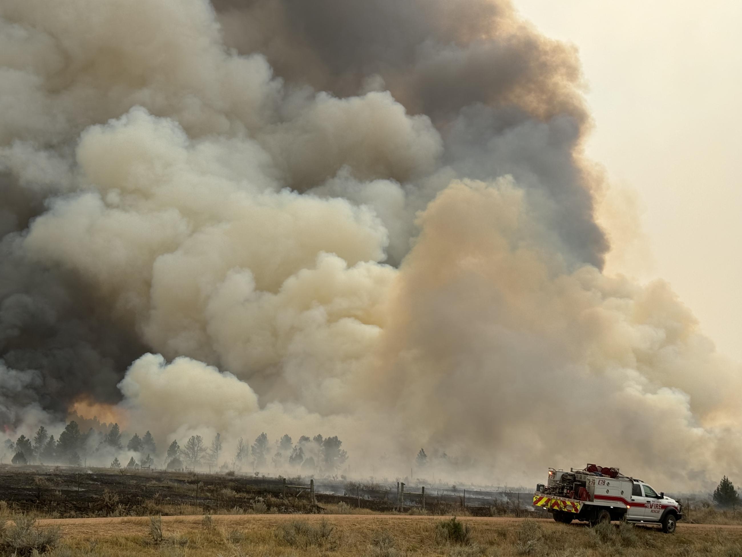 A plume of smoke billows in the background of a fire engine in the foreground on short grass.