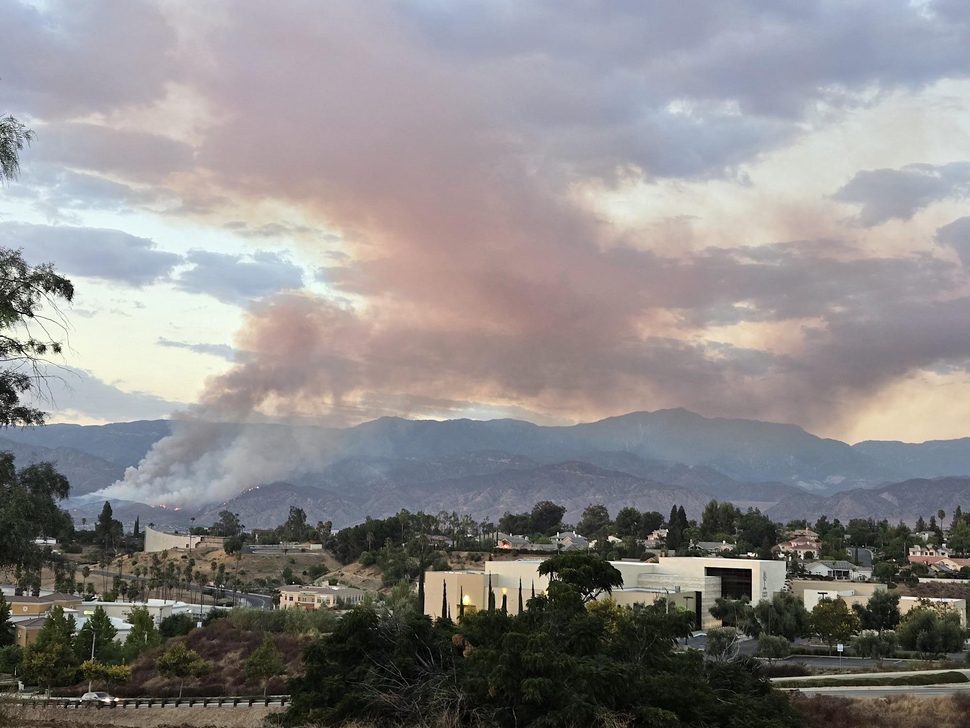 A wide angle shot showing the wide smoke column of the Line Fire. Mountains are in the background with a partly cloudy sky. The valley is at the bottom of the photograph. 