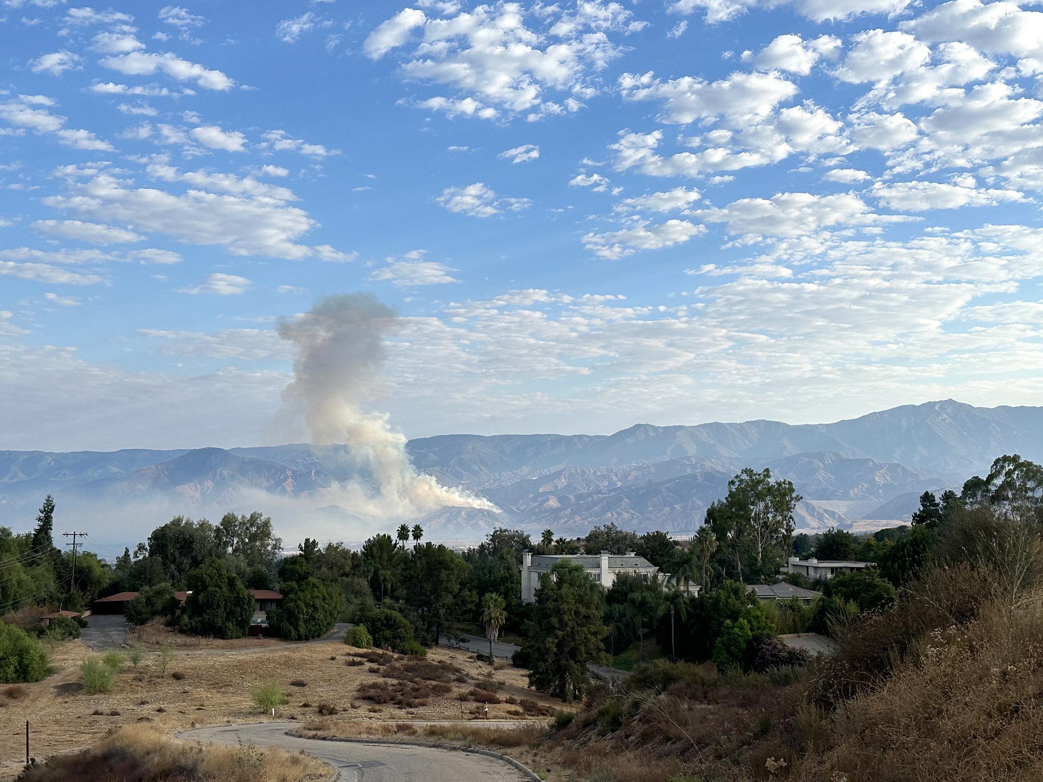 A far away shot of the Line Fire smoke column, rising slightly left on the photograph. Sky is half filled with fluffy white clouds, mountain are in the background and a line of green trees fill the bottom of the photo. 