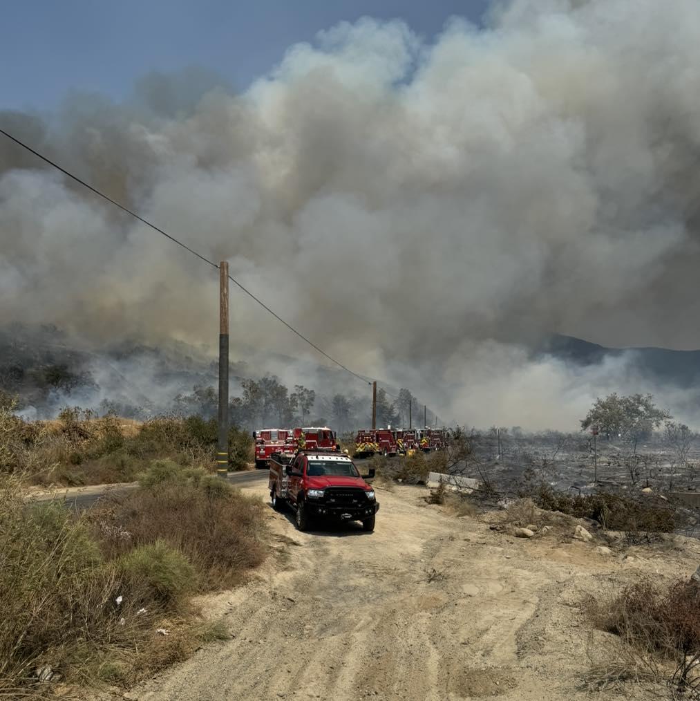 Fire engines turn on to a dirt road while working on the fire. Billowing dark grey smoke fills the photo behind them entirely.