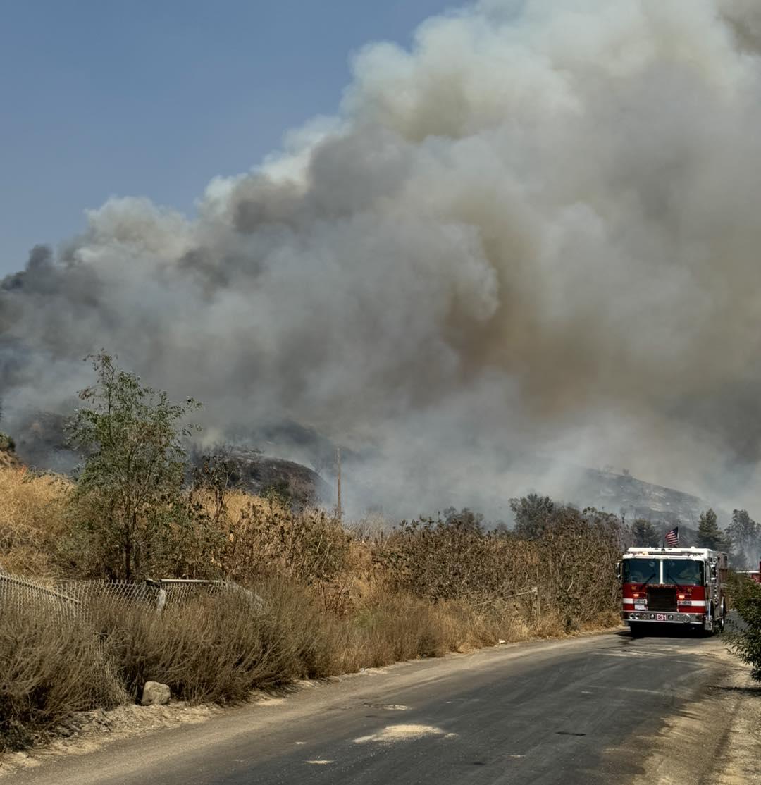 A road with a large red fire engine is in the lower left side of the photo. Brushy vegetation is along the road on the left side and thick dark grey smoke billows from the vegetation over towards the engine.