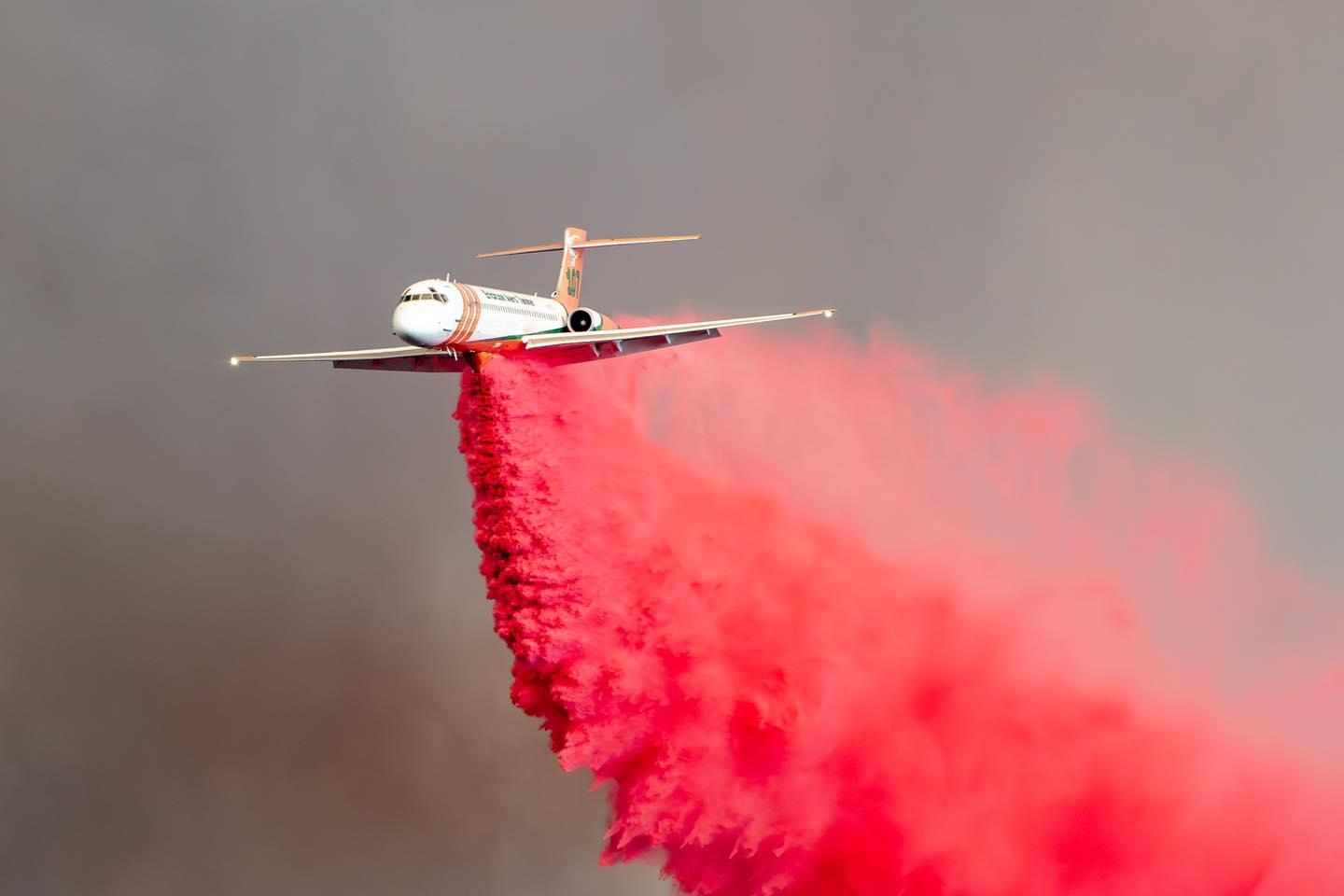 A very large air tanker is in the center of the photo with red fire retardant being dispersed from under it. The sky is completely filled with dark grey smoke.