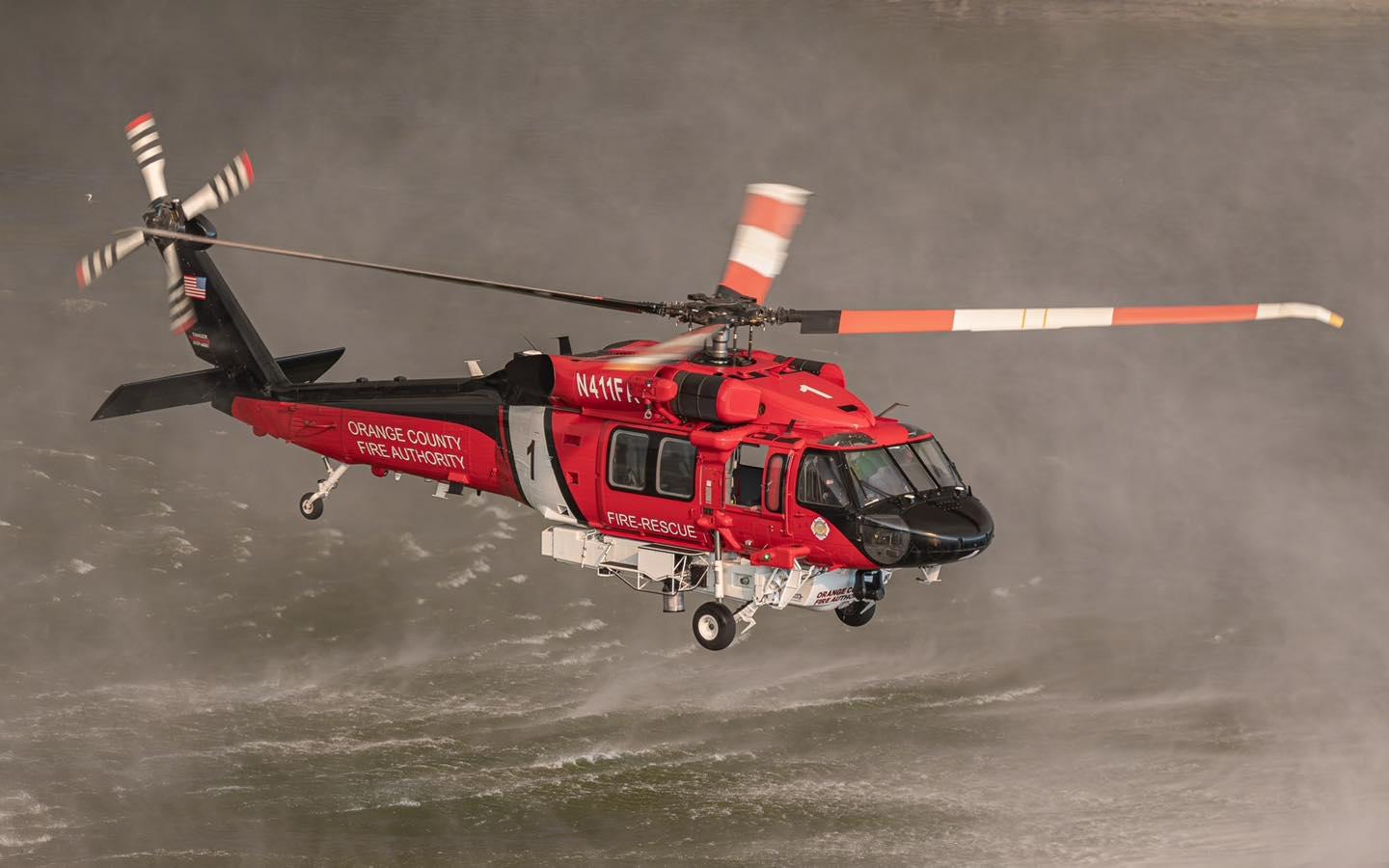 A mainly red helicopter is lowering itself on to a body of water. It will fill up with water. The water is wavy from the wake of the helicopter rotors.