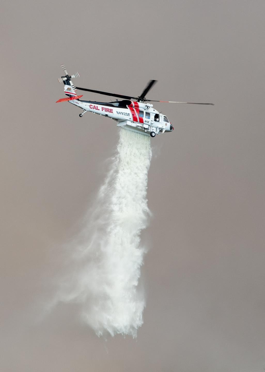 The sky is totally dark grey and fills the photo. A helicopter is in center, releasing water on to the fire. The water is coming from the body of the helicopter.