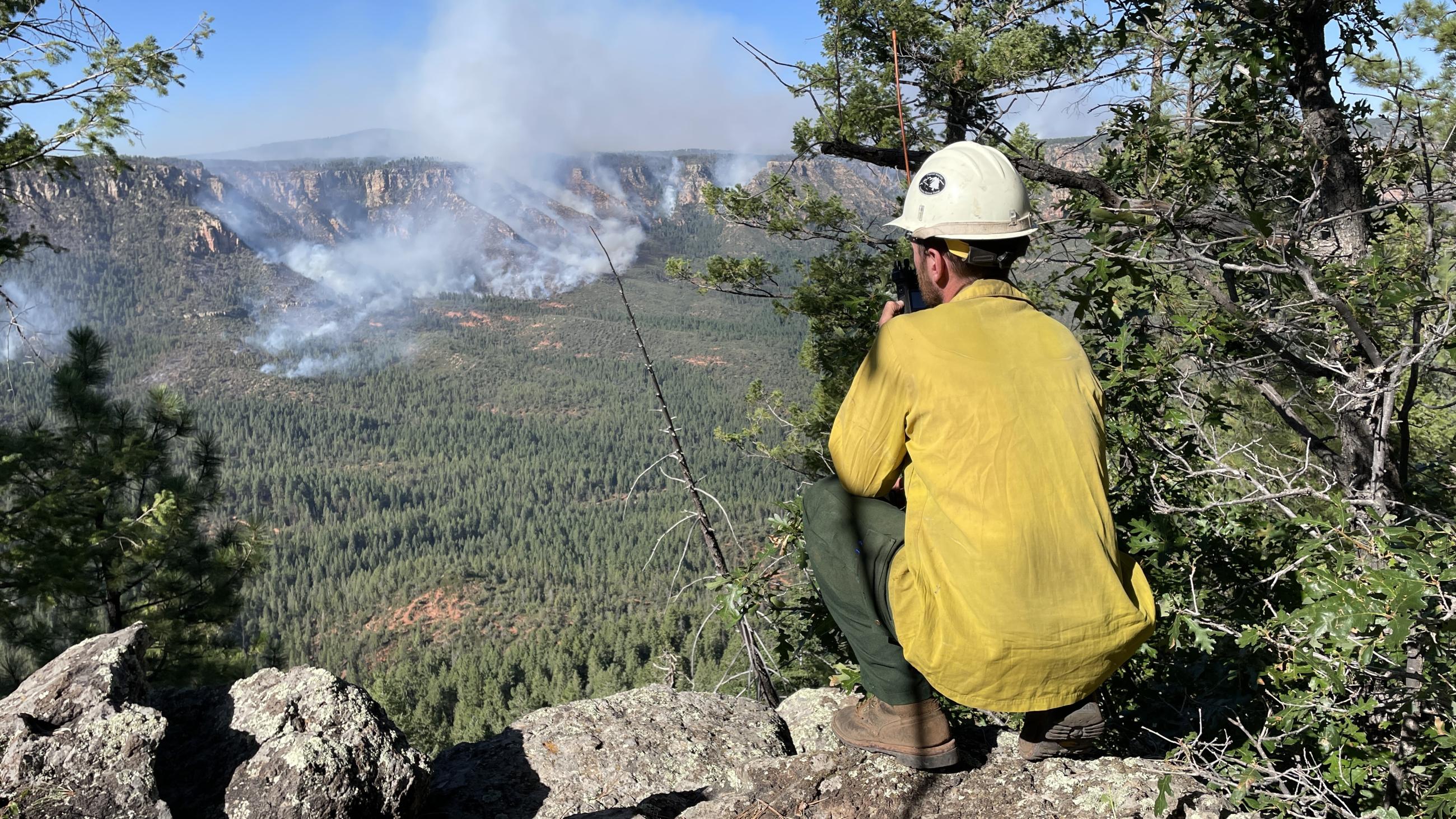 Man in yellow shirt and hardhat crouched down with a radio looks over a valley with smoke in the distance.