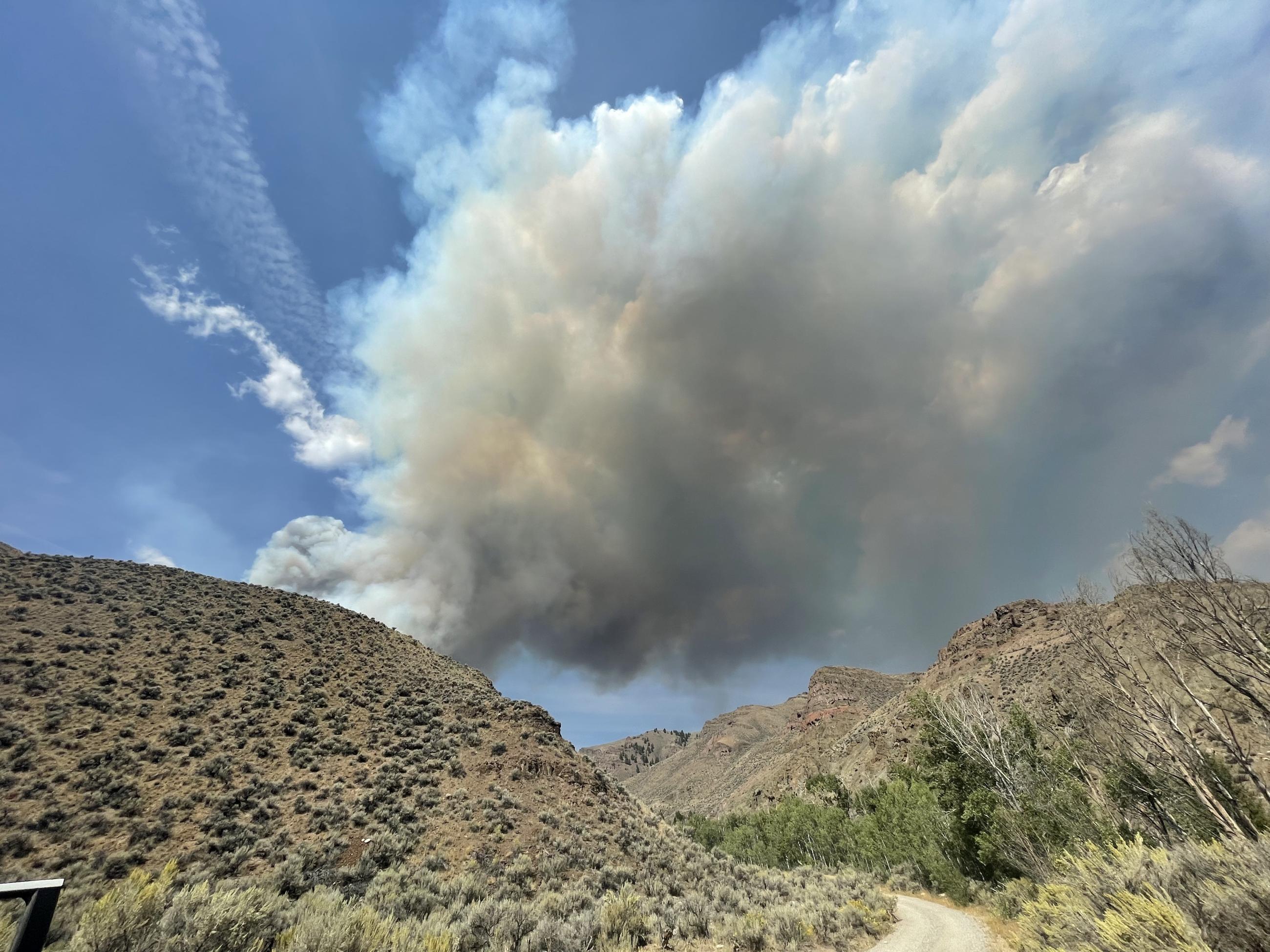 Dirt road winds between sage-covered hills, leading to a plume of smoke that billows to the top left of the landscape with a bright blue sky beyond.