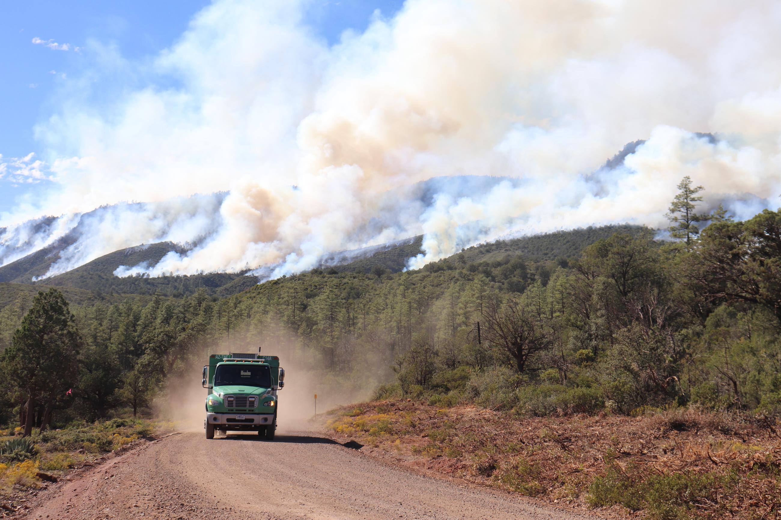 Green fire truck driving down a dirt road with smoke on mountains in background. 