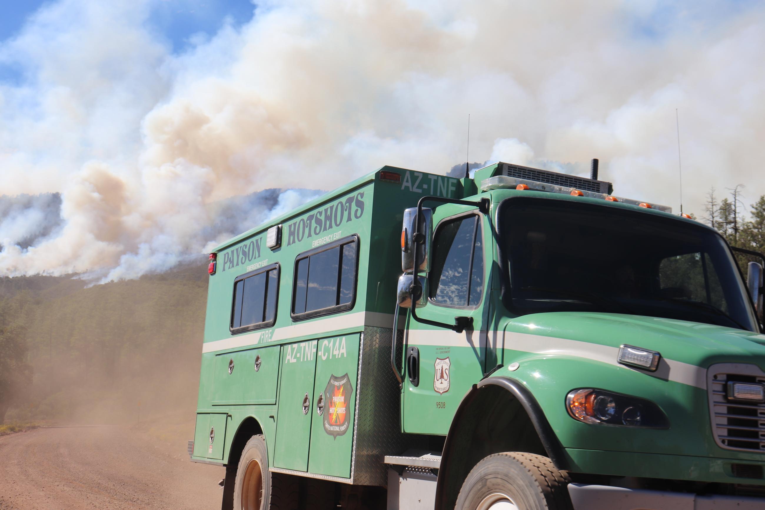 A green fire truck driving down a dirt road with smoke on the mountains behind.