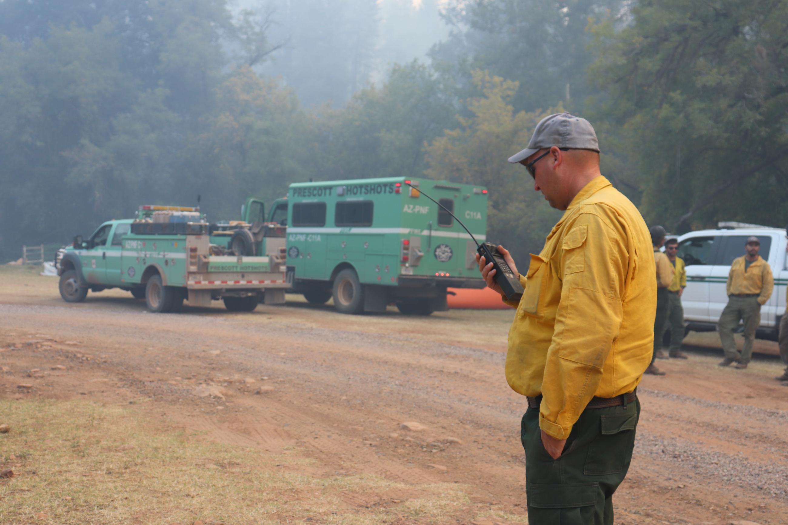 man in yellow shirt talks on a radio with crew in background and green trucks.