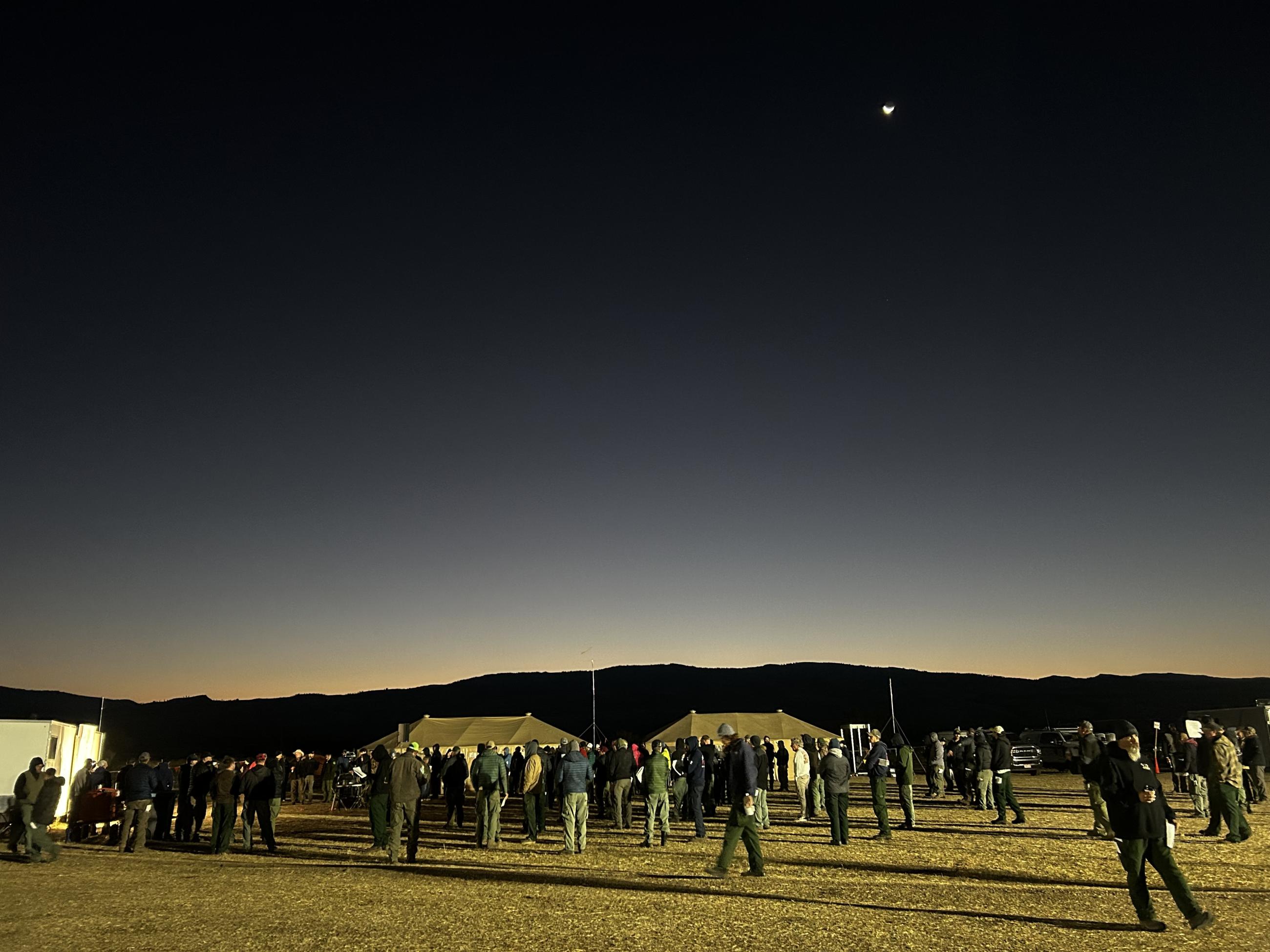 wide angle photo of people gathered at morning briefing as the sun begins to rise over mountains in the background