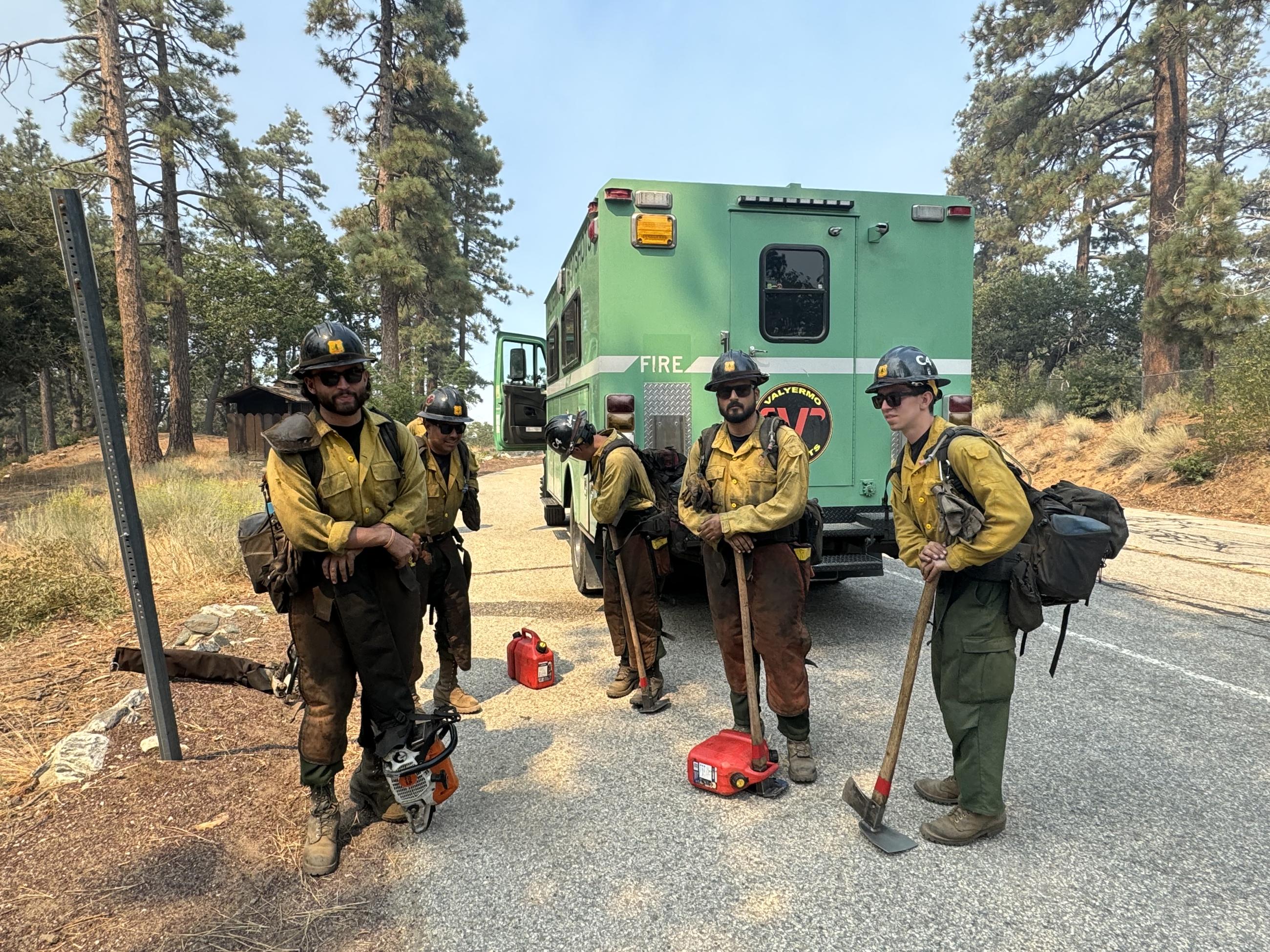 Four hotshots in yellow and green uniforms in front of a green fire truck with tools