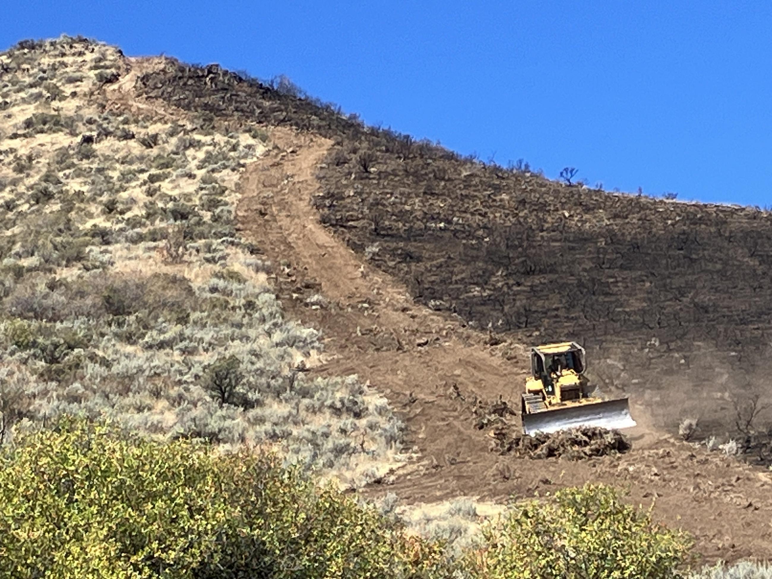 A dozer creating a direct fire line down a hill with burnt sagebrush and grass to the right of the line and unburnt sagebrush and grass to the left