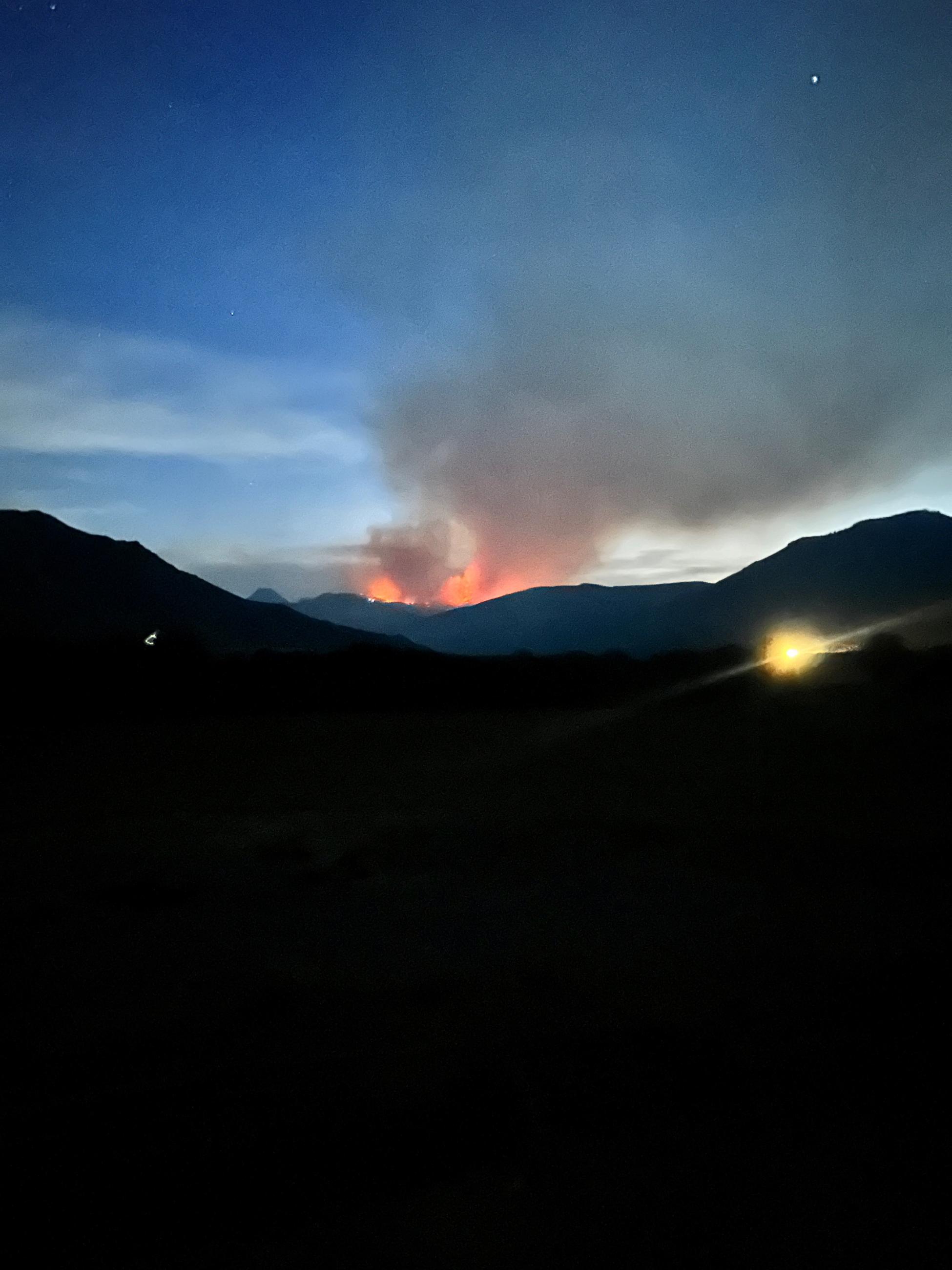 An orange glow is seen over dark mountains with stars in the dark blue sky