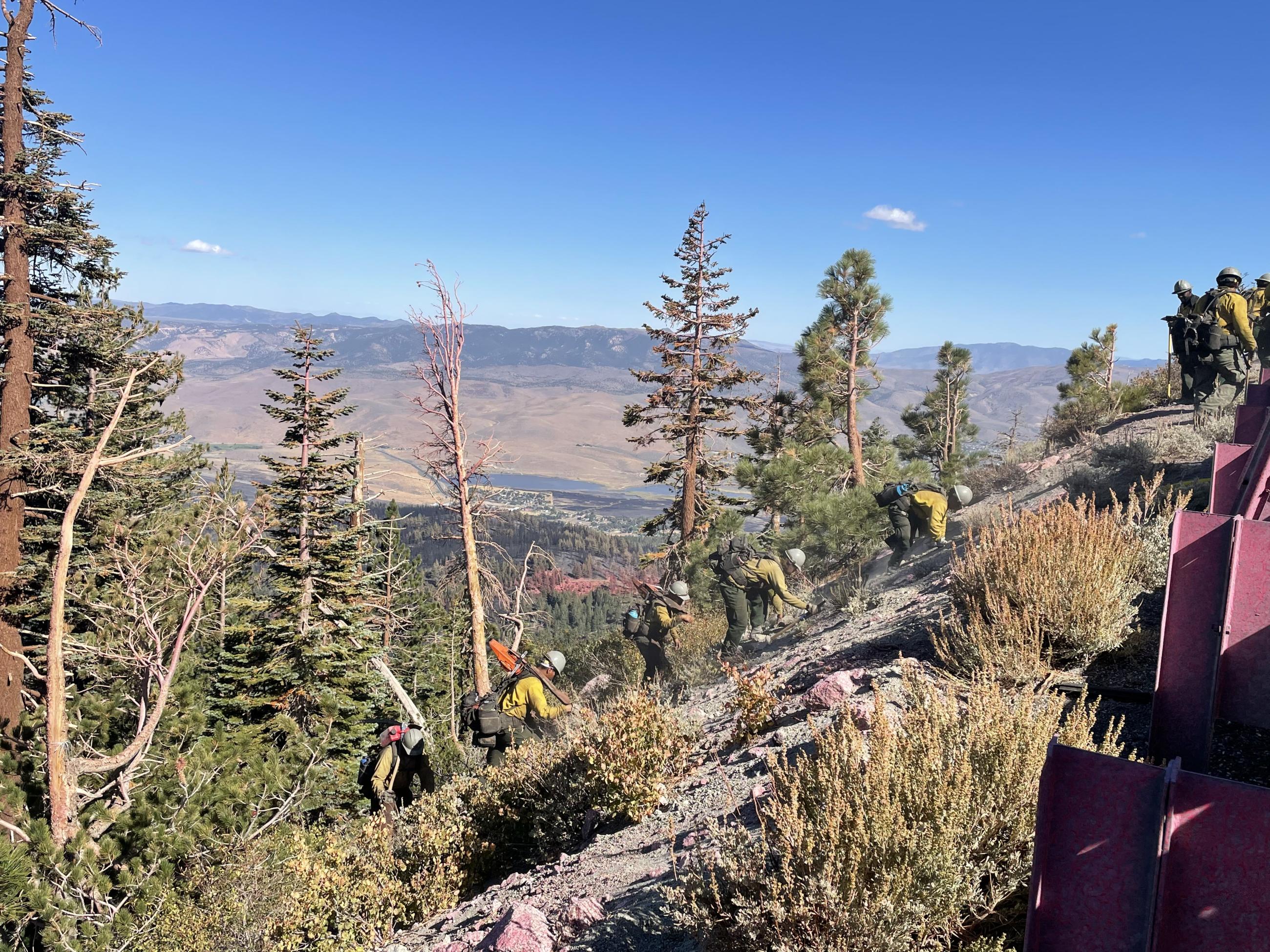 Photo showing five firefighters climbing steep hillside, two carrying chainsaws.  Washoe Valley visible behind them.