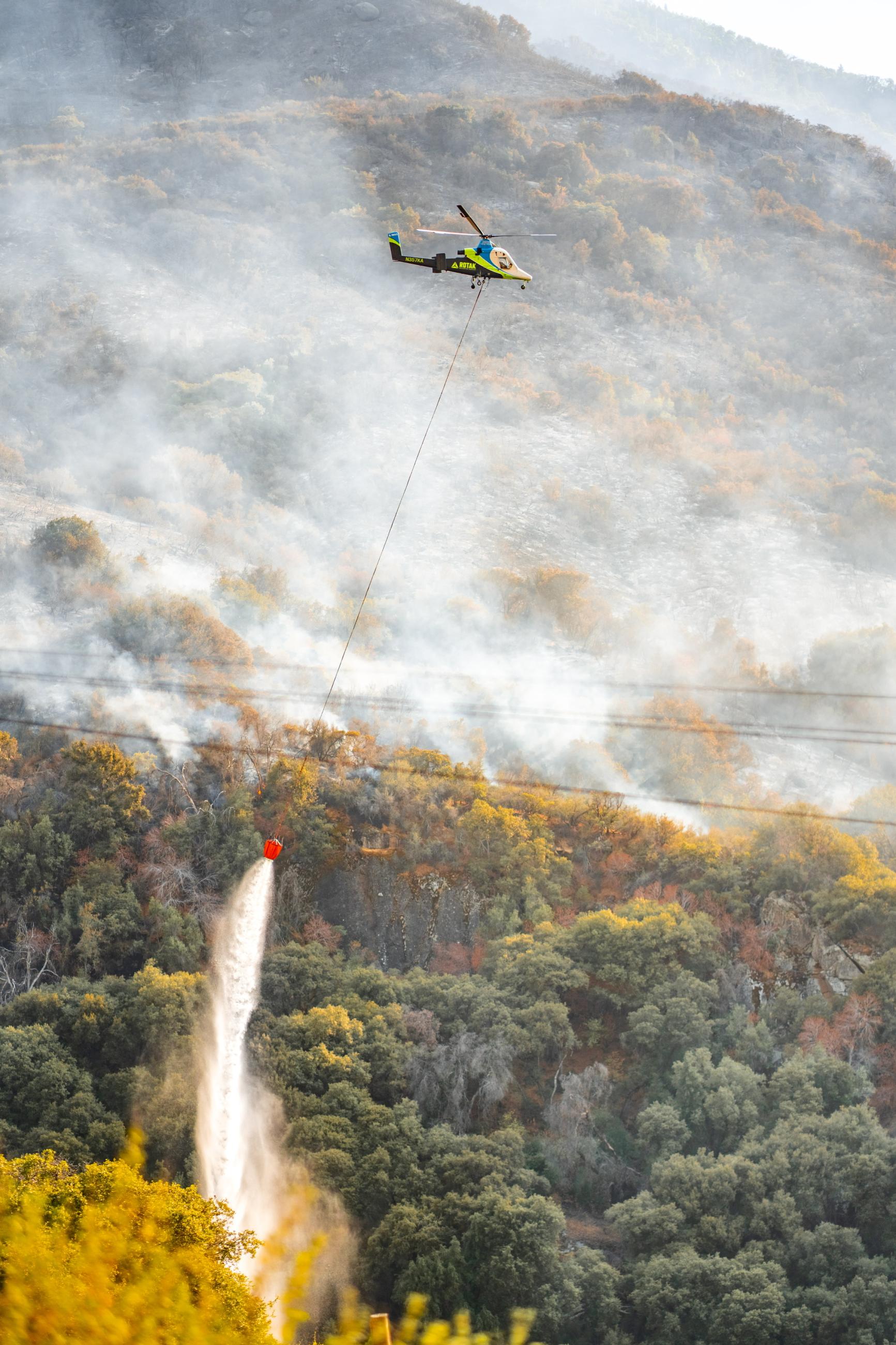 Water streams out of a large orange bucket suspended by a long line underneath a large helicopter as it flies over a smokey mountainside.