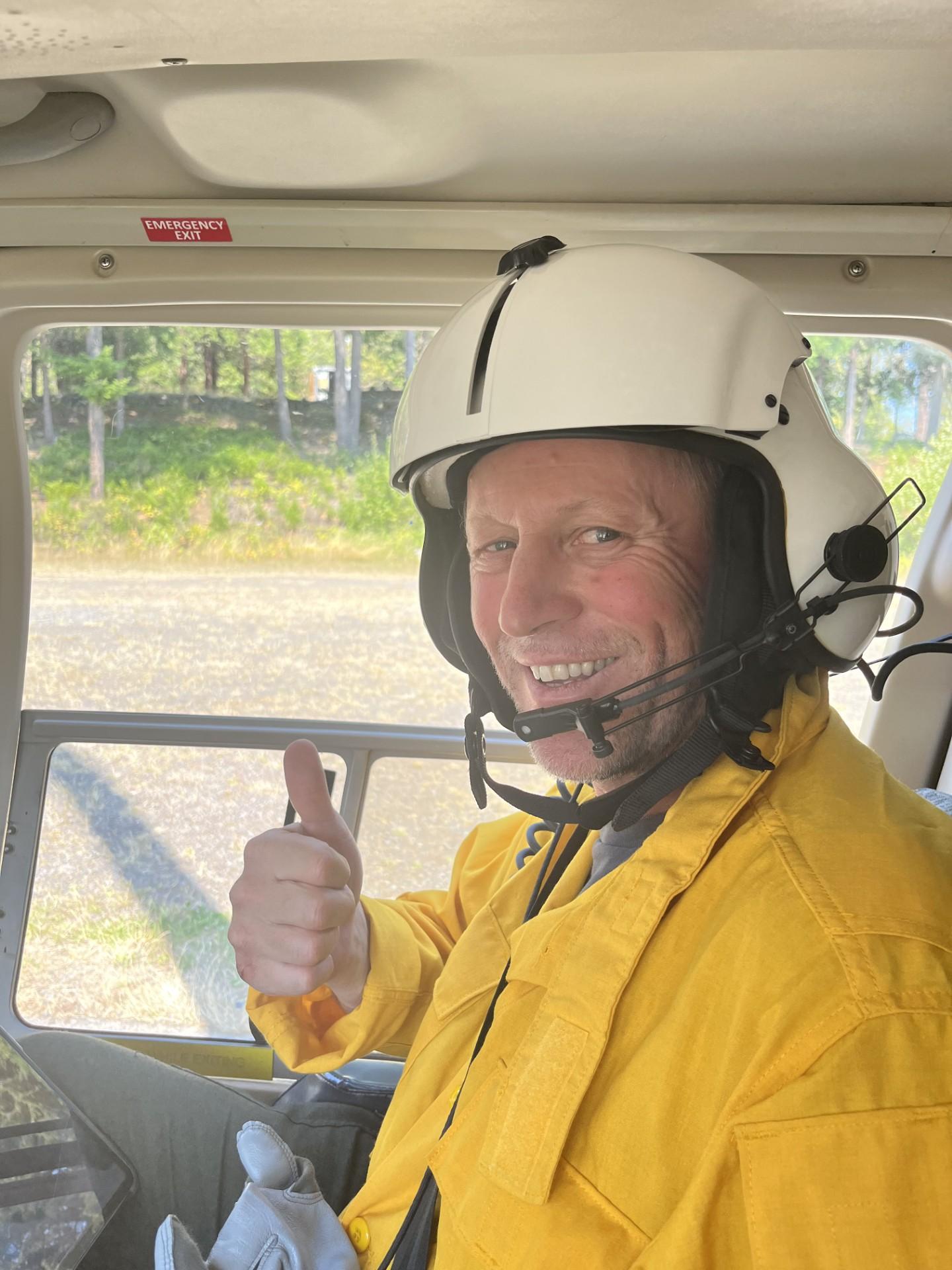 Fire Behavior Analyst Aleks Maric in yellow shirt and helmet gets ready for helicopter flight with a thumbs up