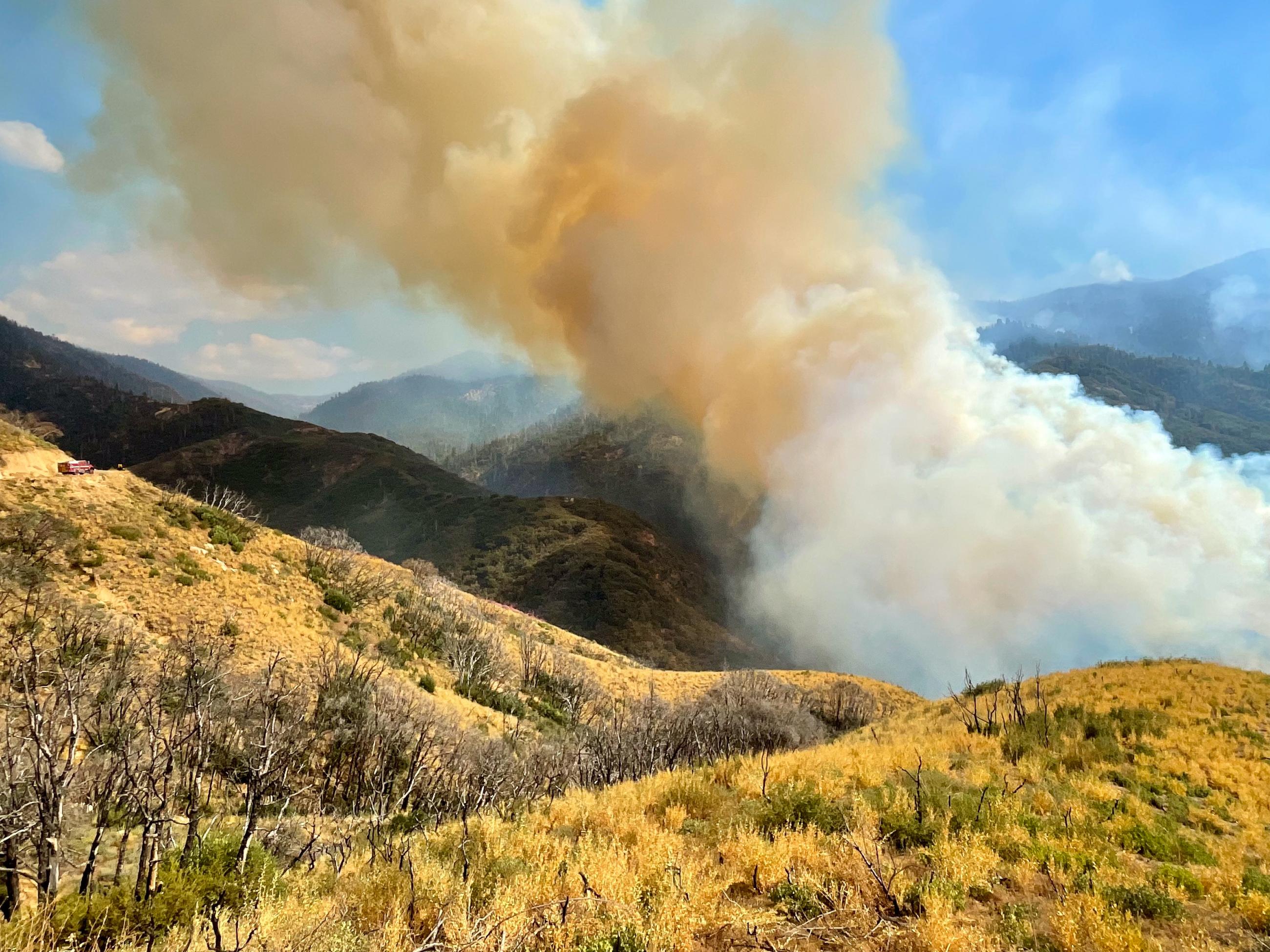 Smoke rises off of a hill and mountain landscape. 