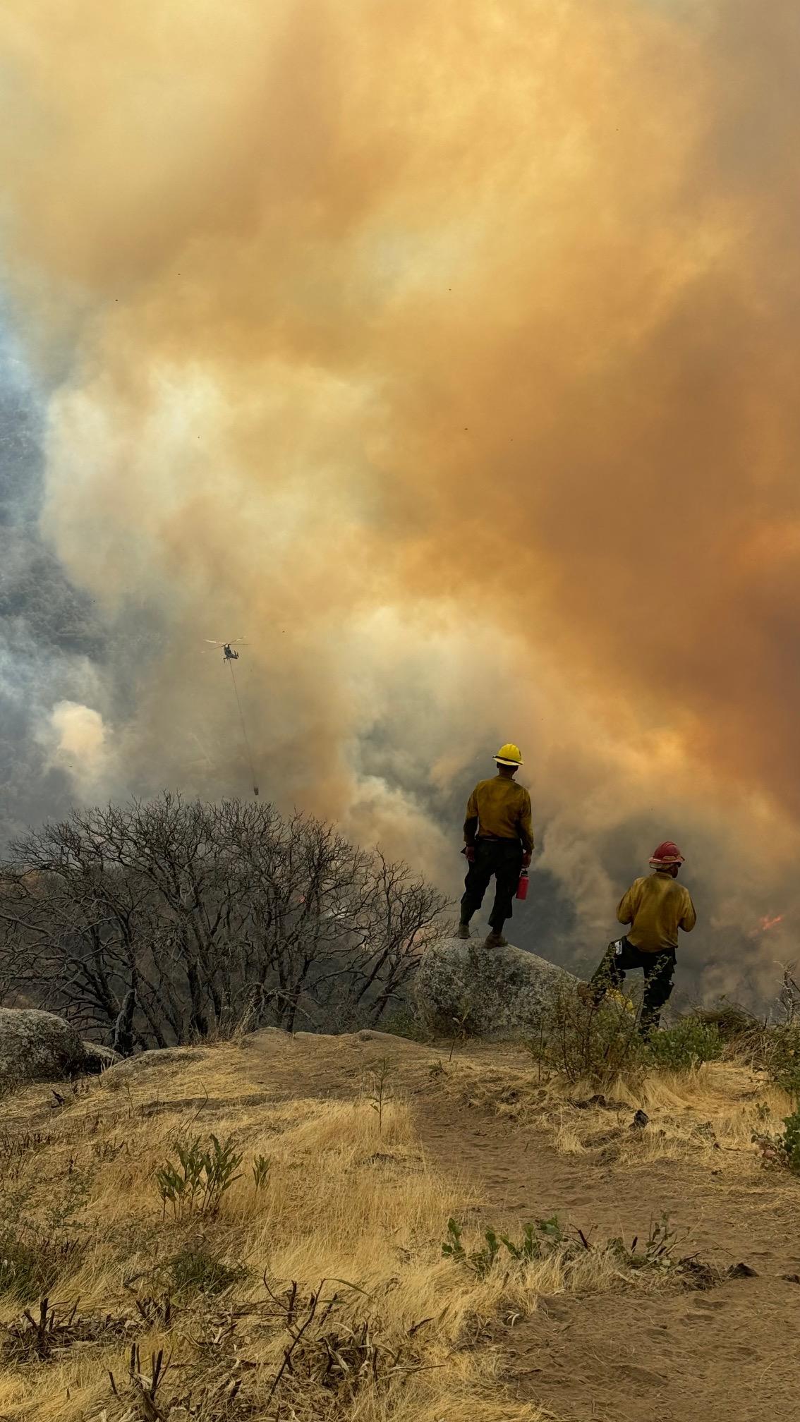 Two firefighters look at smoke in a valley and a helicopter