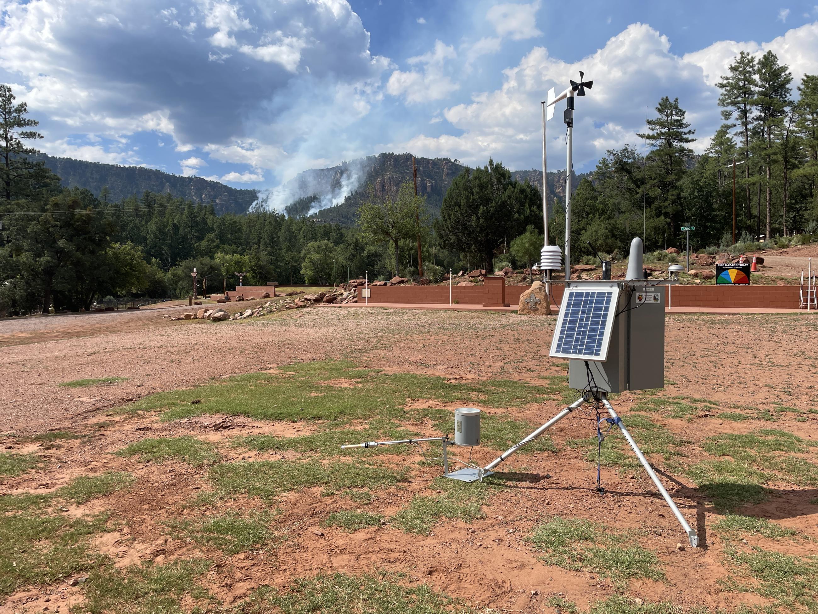 a grey metal box with solar panel and cables in a dirt lot with smoke in the background