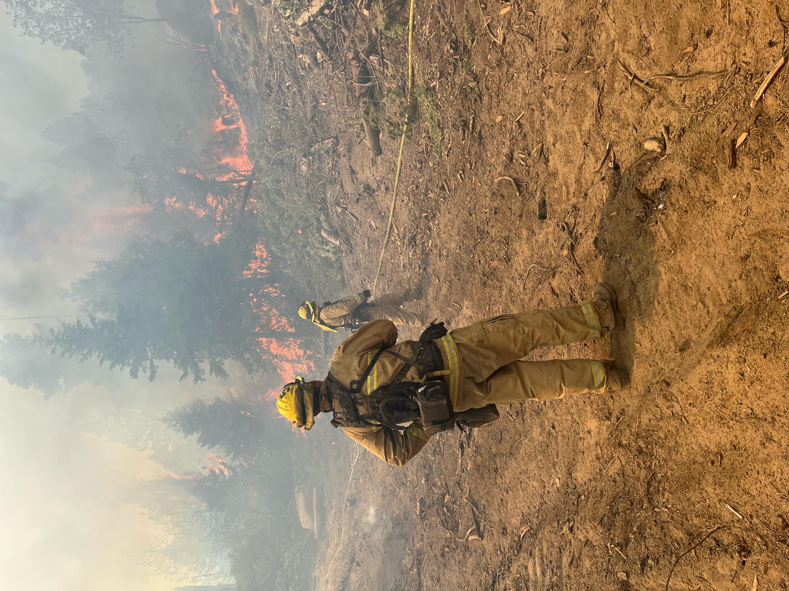 Firefighters and a hose on the ground with flames in the background.