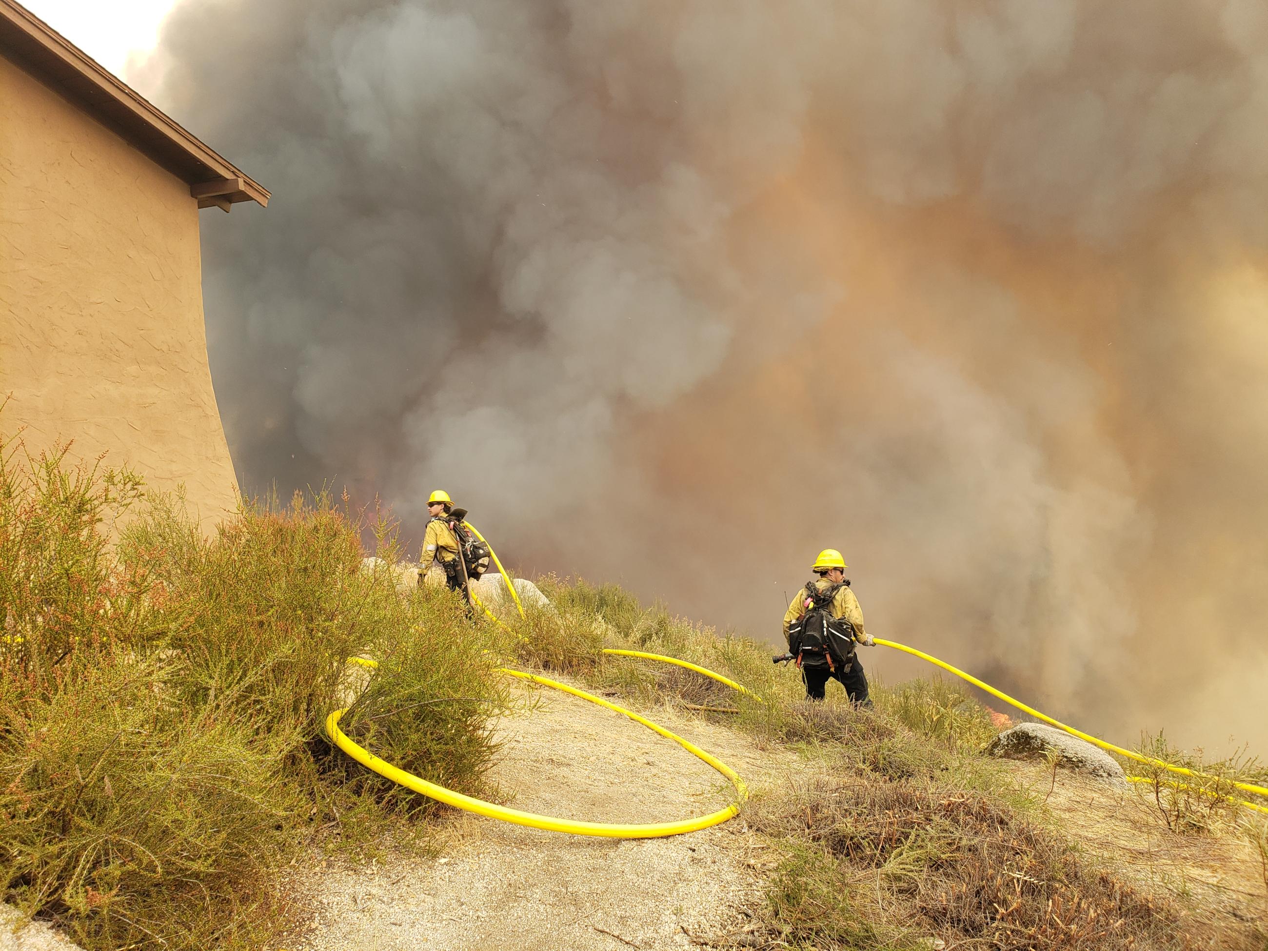 Two firefighters and a hose next to a building with smoke in the background. 