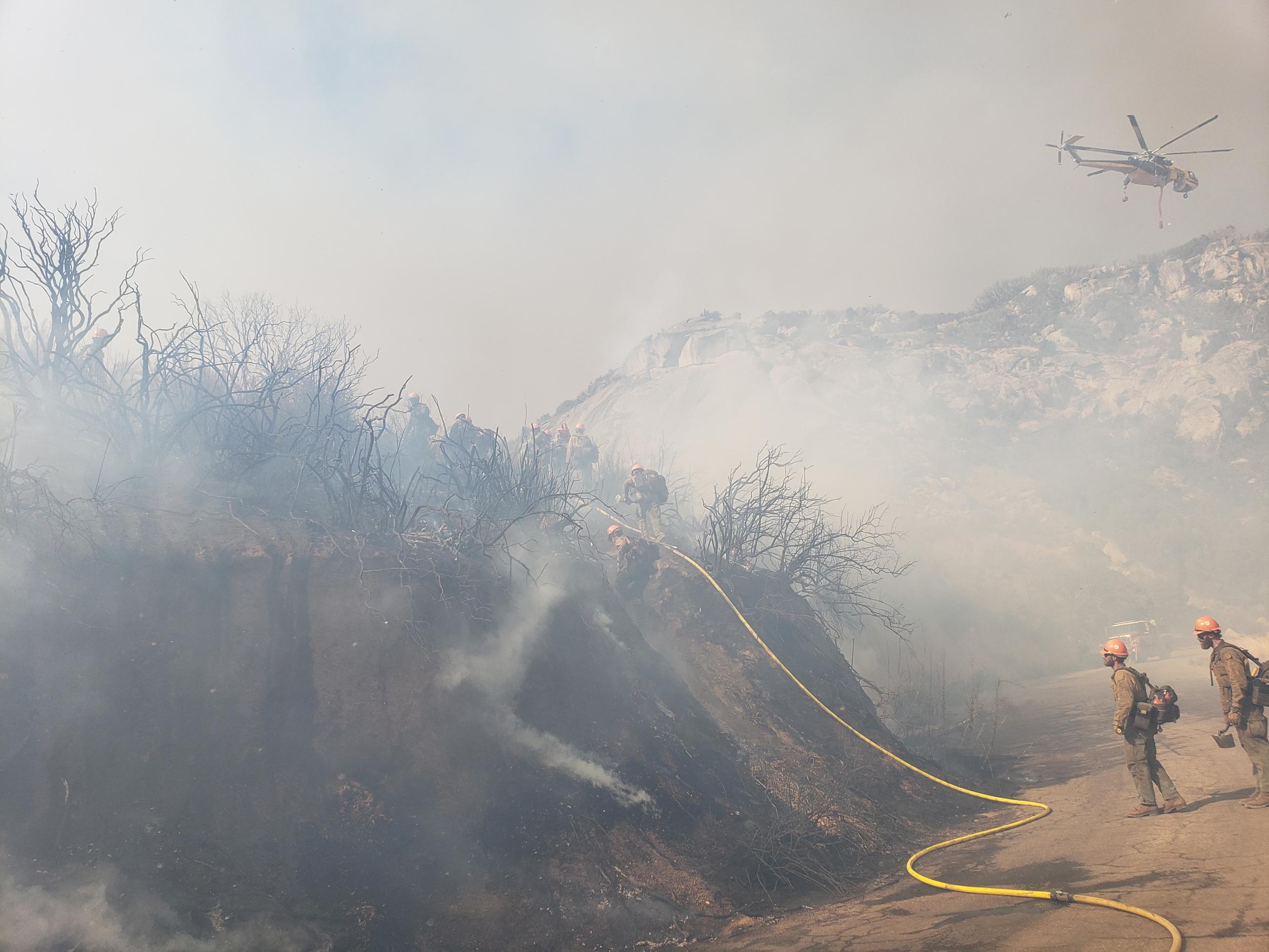 Firefighters carrying a hose across a dirt road, a helicopter is in the air in the upper right corner.