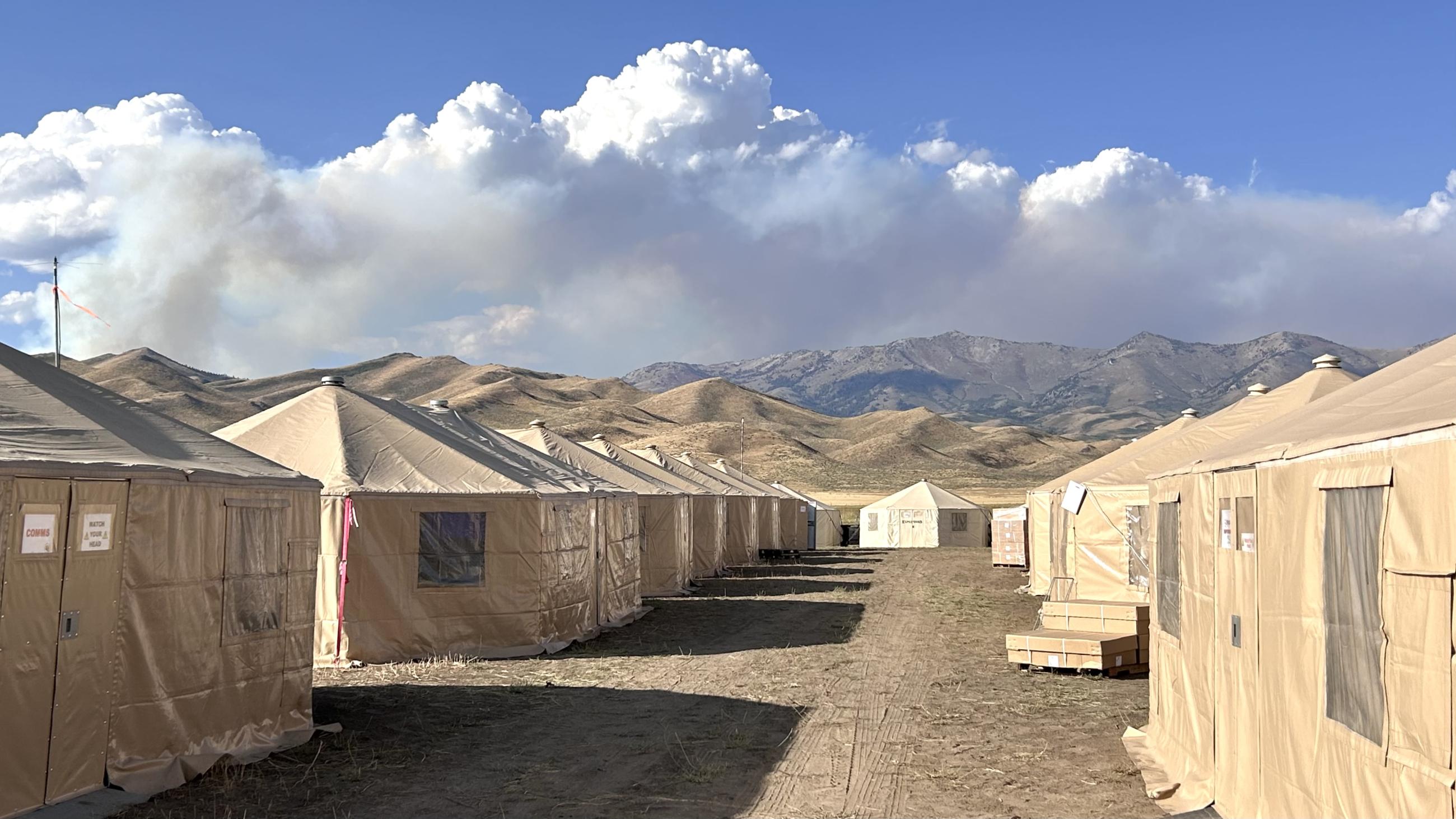 White yurts on dirt field with Chimney Fire smoke in the background above ridge.