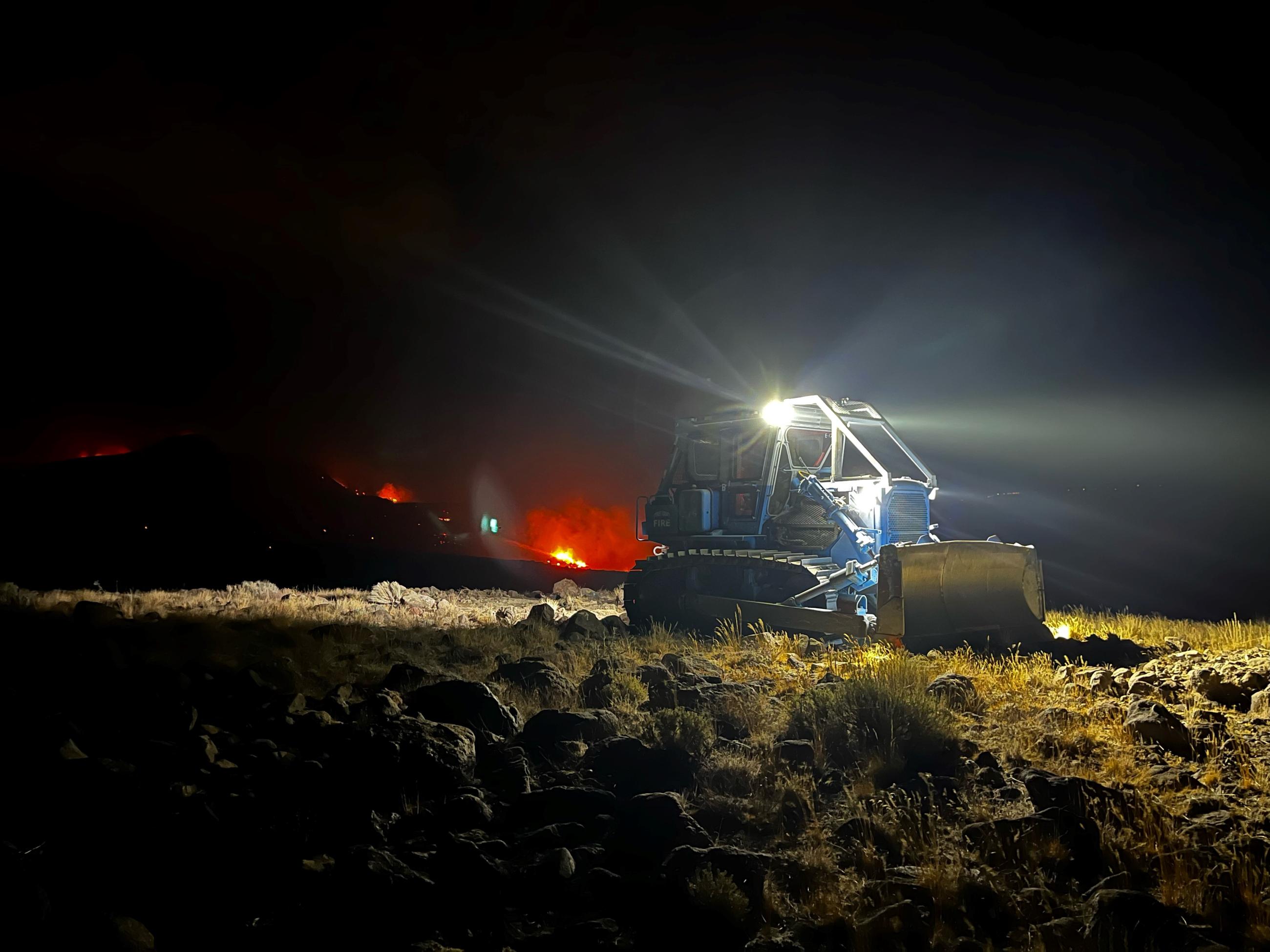 Photo of bulldozer at night with fire behind it.