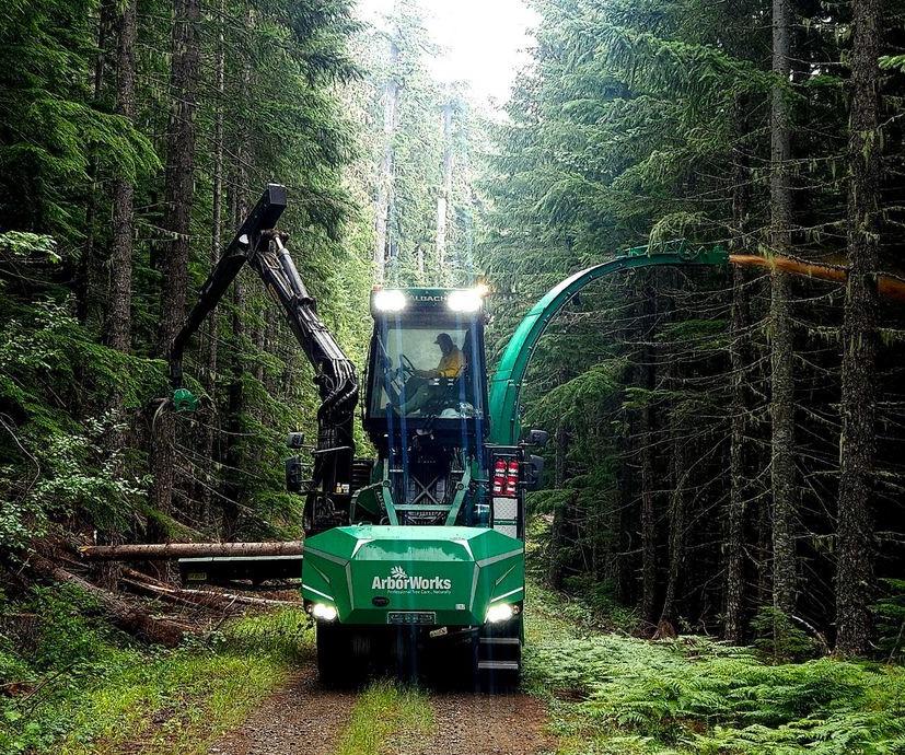 A green vehicle with lights on top, a crane arm to the left moving the logs and a metal to the right shooting wood chips. The vehicle is on a dirt road surrounded by a conifur forest.