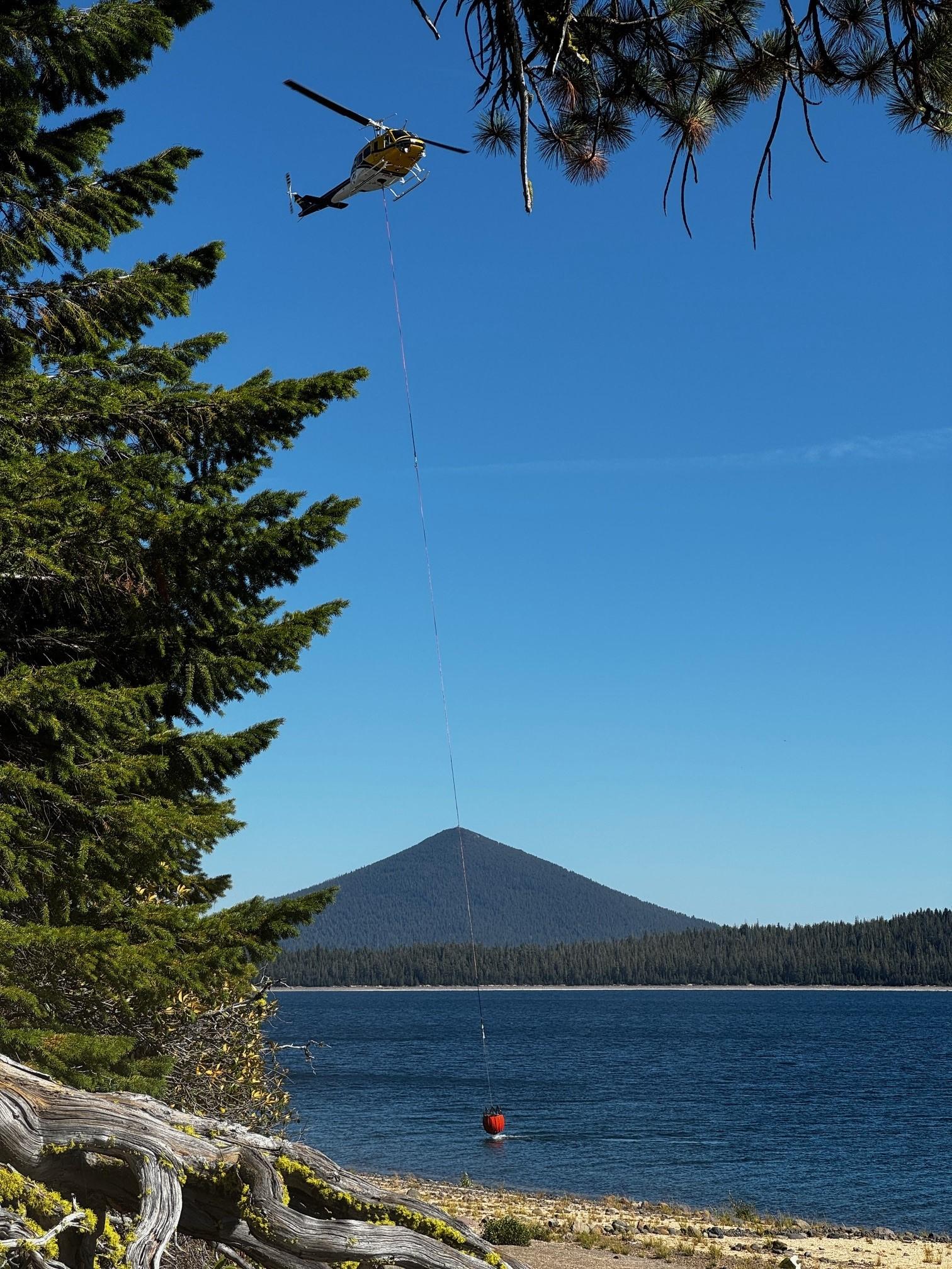 Helicopter hovering over lake with a water bucket underneath as they lower to fill the bucket