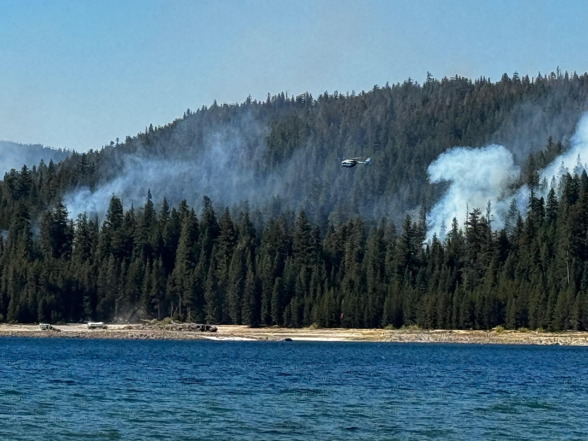 A helicopter is flying over a wildfire with a lake in the foreground and forest in the background