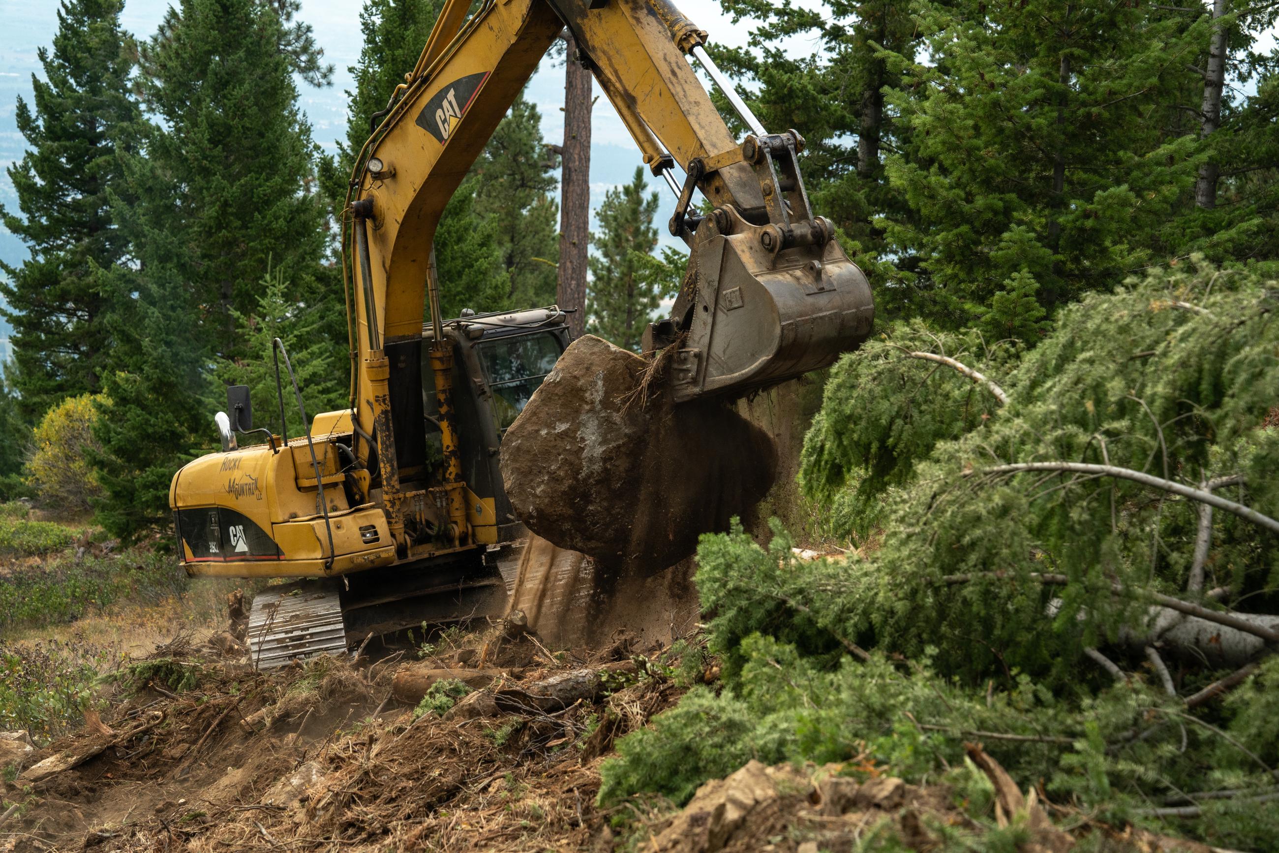 A yellow excavator drops a large rock as dirt spills out of the scoop while doing suppression repair work on the Sharrott Creek Fire. 