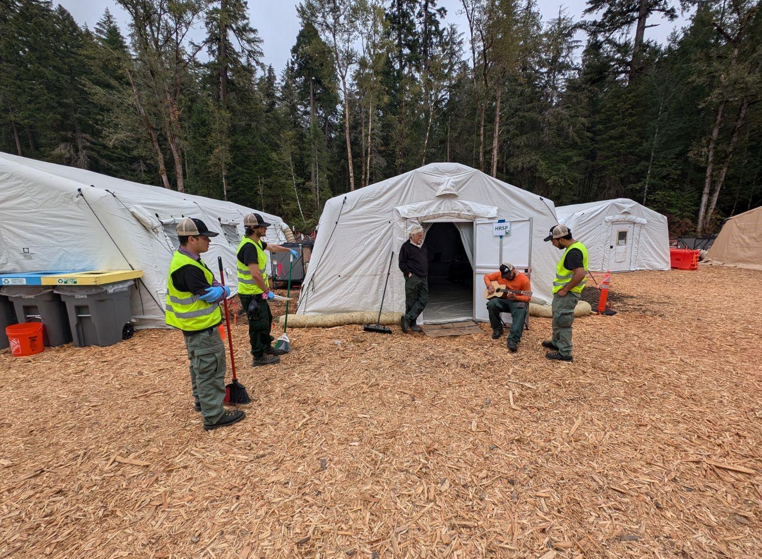 In front of a large white tent sits a young many playing guitar. To the right stands a man in a yellow vest. To the left, stand two men holding brooms and a dust pan.