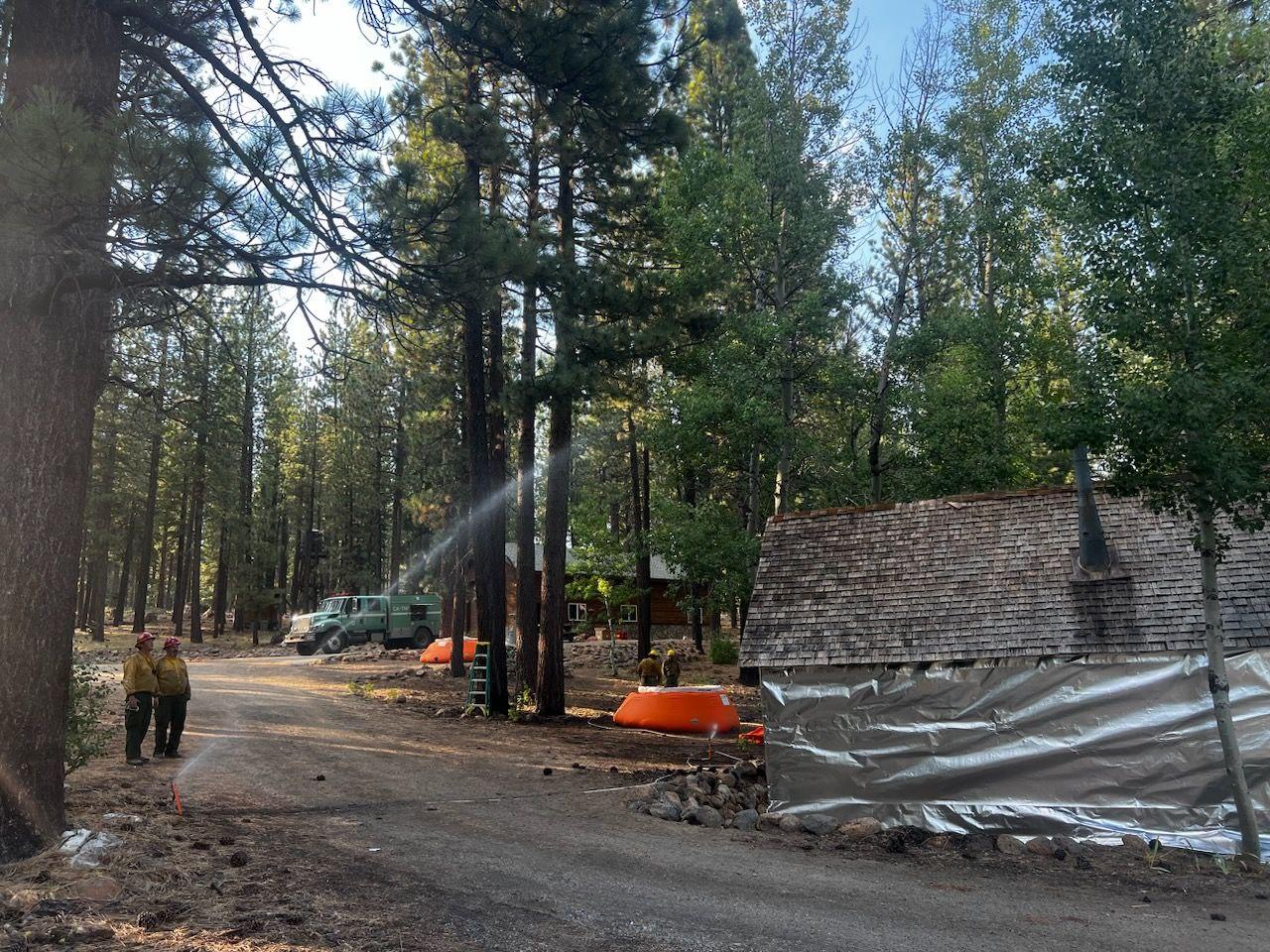 Photo of forest with a structure that is wrapped for a fire. There is a firefighter with a hose spraying the structure.