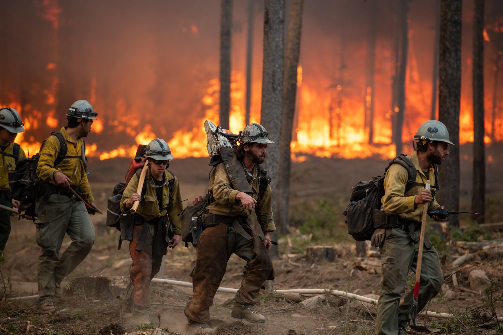 Four Firefighters hiking with gear in front of forest trees and flames