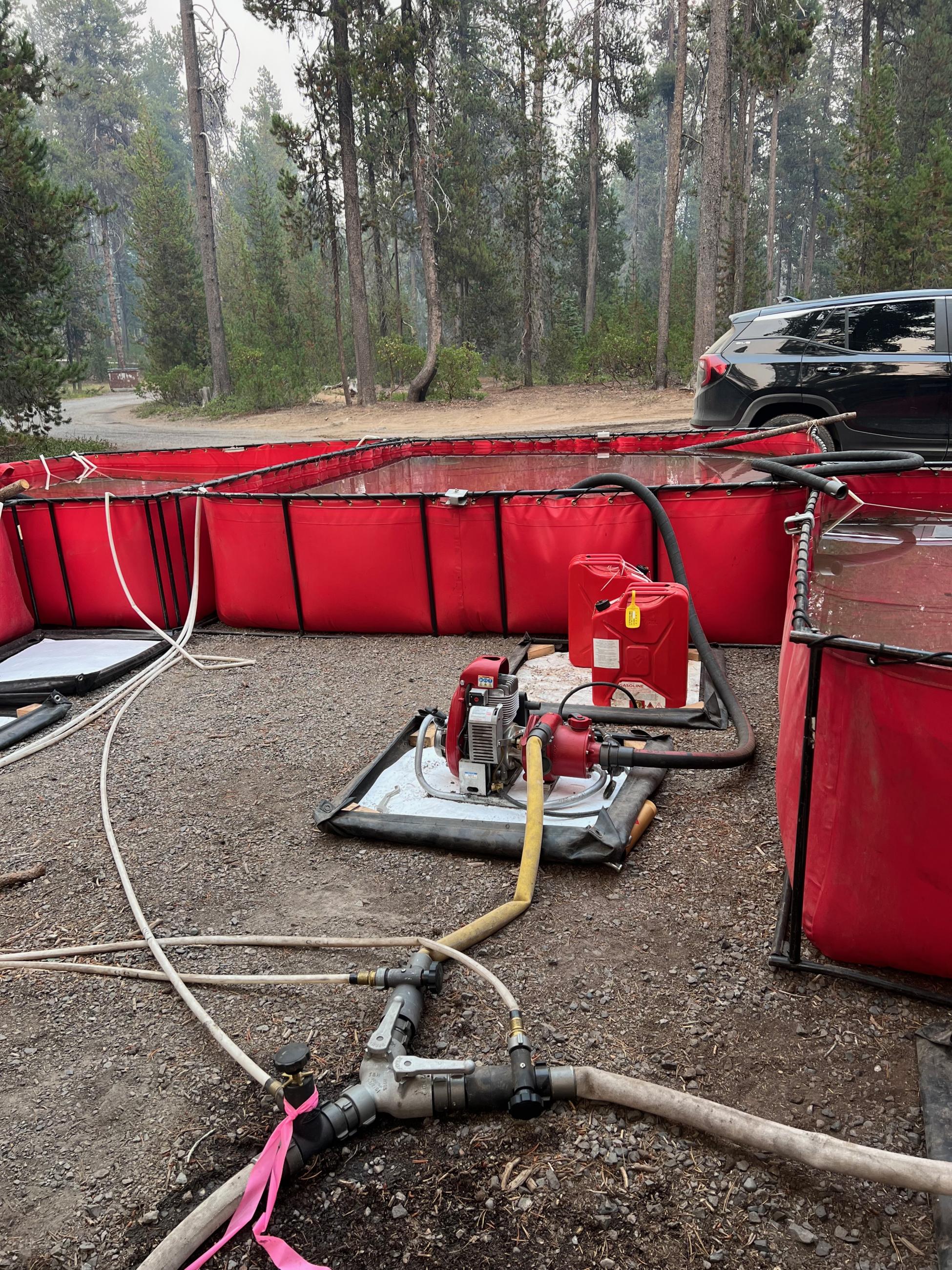 A water pump with hoses coming off it is in the foreground with red portable water tanks beyond.