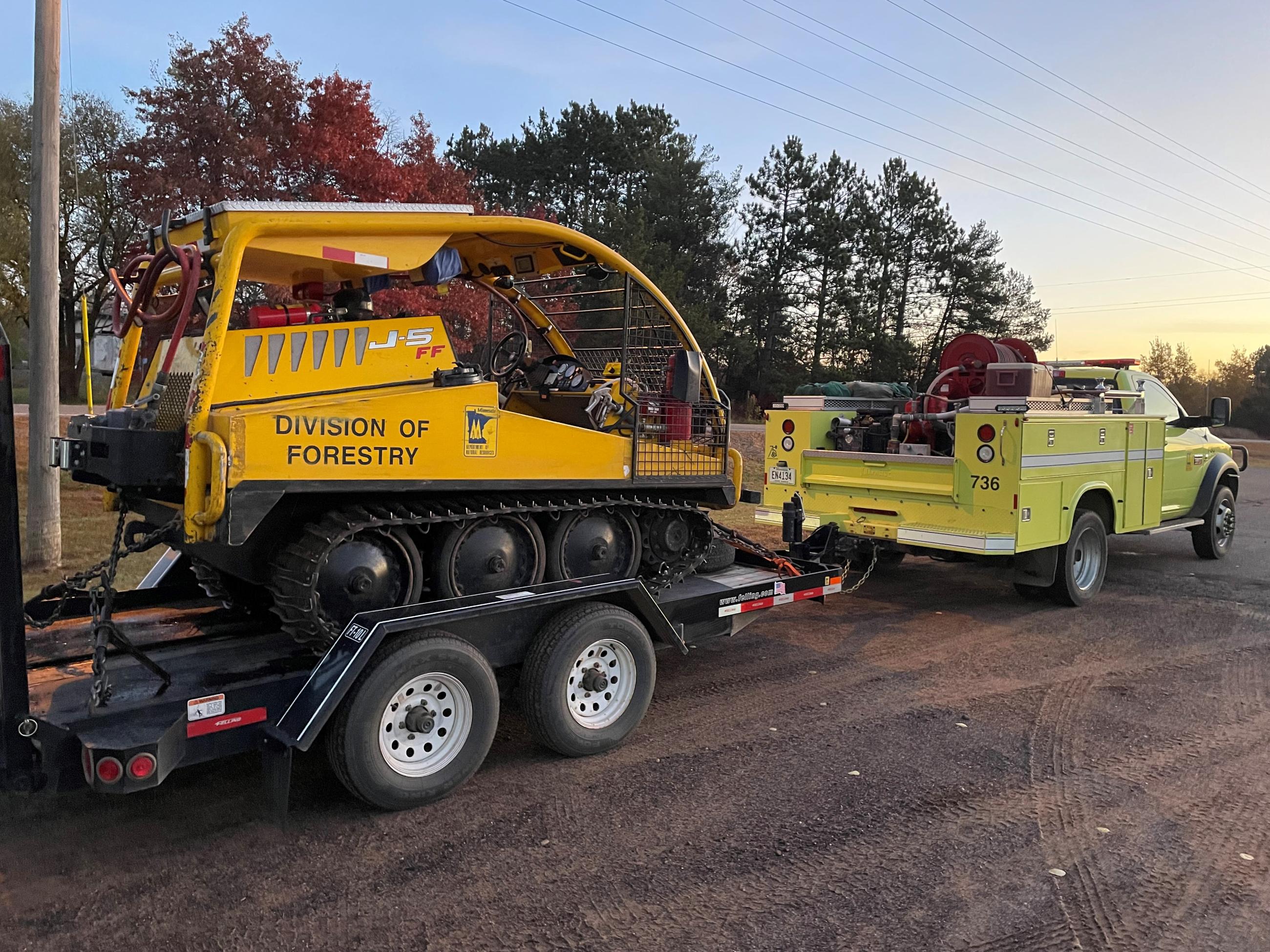 Image of a yellow tracked vehicle on a trailer attached to a fire truck. 